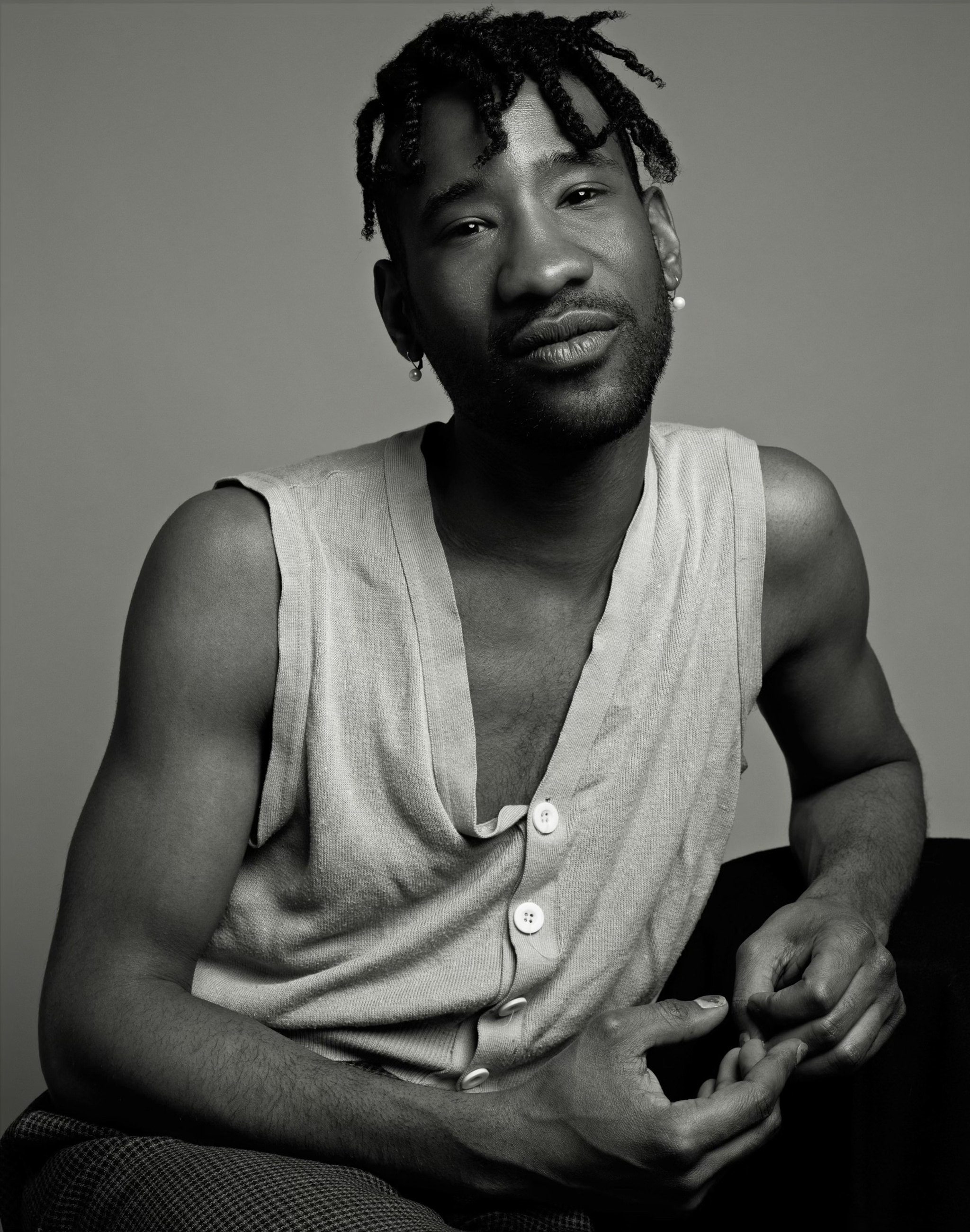 A black-and-white portrait of artist Jarrett Key. Jarrett is a Black person who wears a knit vested buttoned up with no shirt underneath. They lean casually on their left arm, have short locs that fall over their forehead, and look directly at us. 