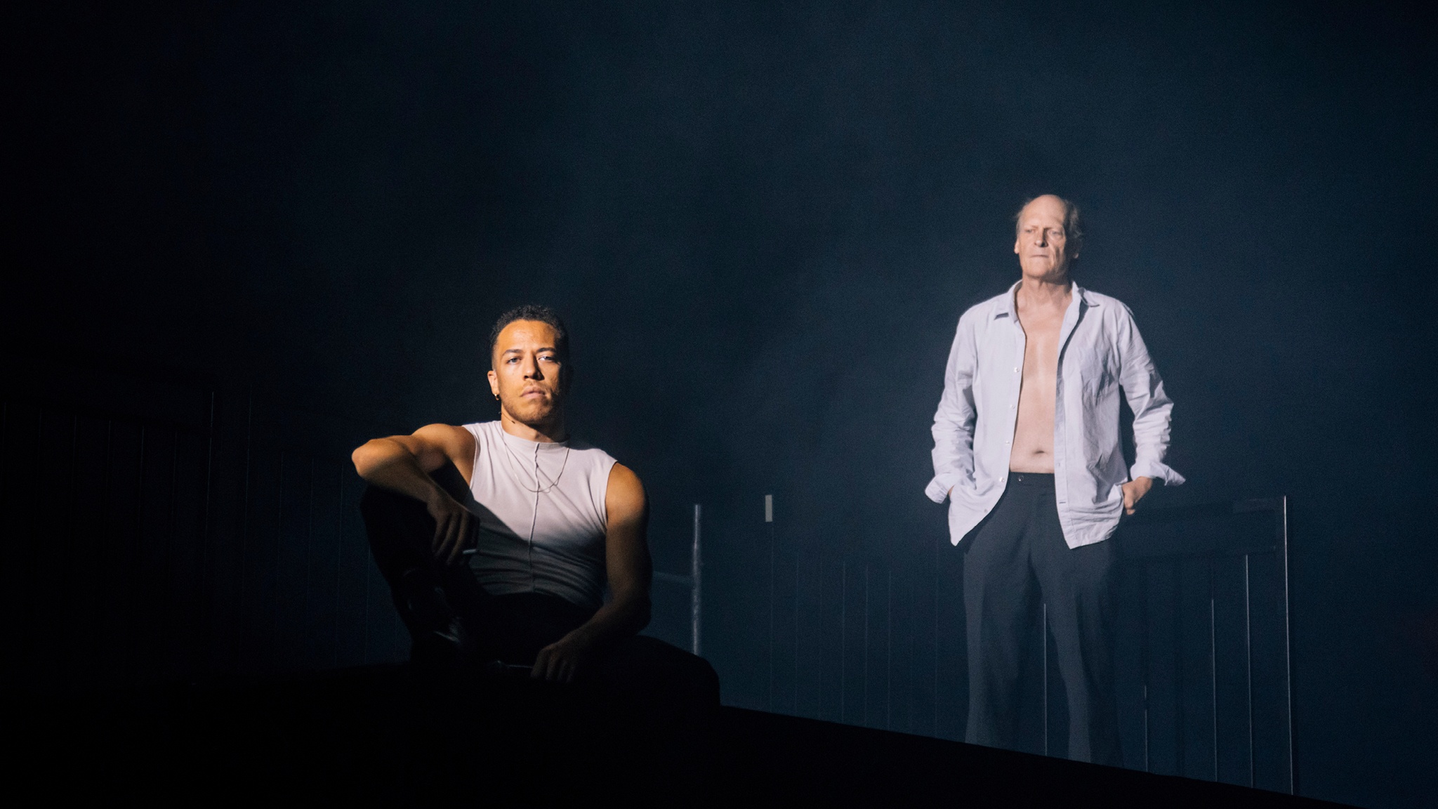 Two performers in Nile Harris's "minor b" stand on a dark stage. They are dramatically lit by a bright light with the rest of the space around them in darkness. One performer, a Black man in a tank top sits with one arm propped on his knee. The other, an older white man in a button down shirt unbuttoned to reveal his bare chest, stands behind him with hands in pockets.