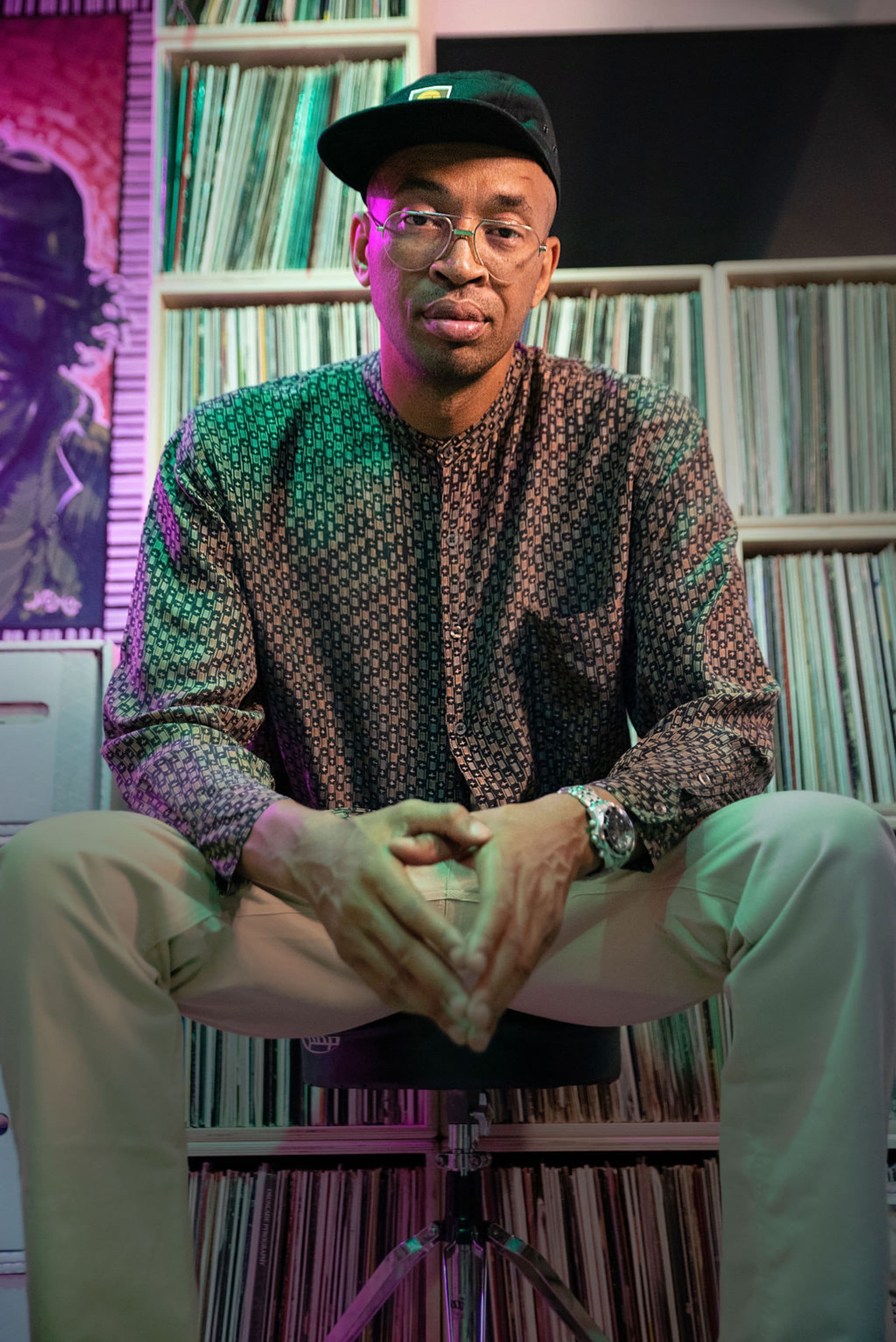 A portrait of DJ Rick Medina sitting with hands between his knees in front of a wall of vinyl records on shelves.