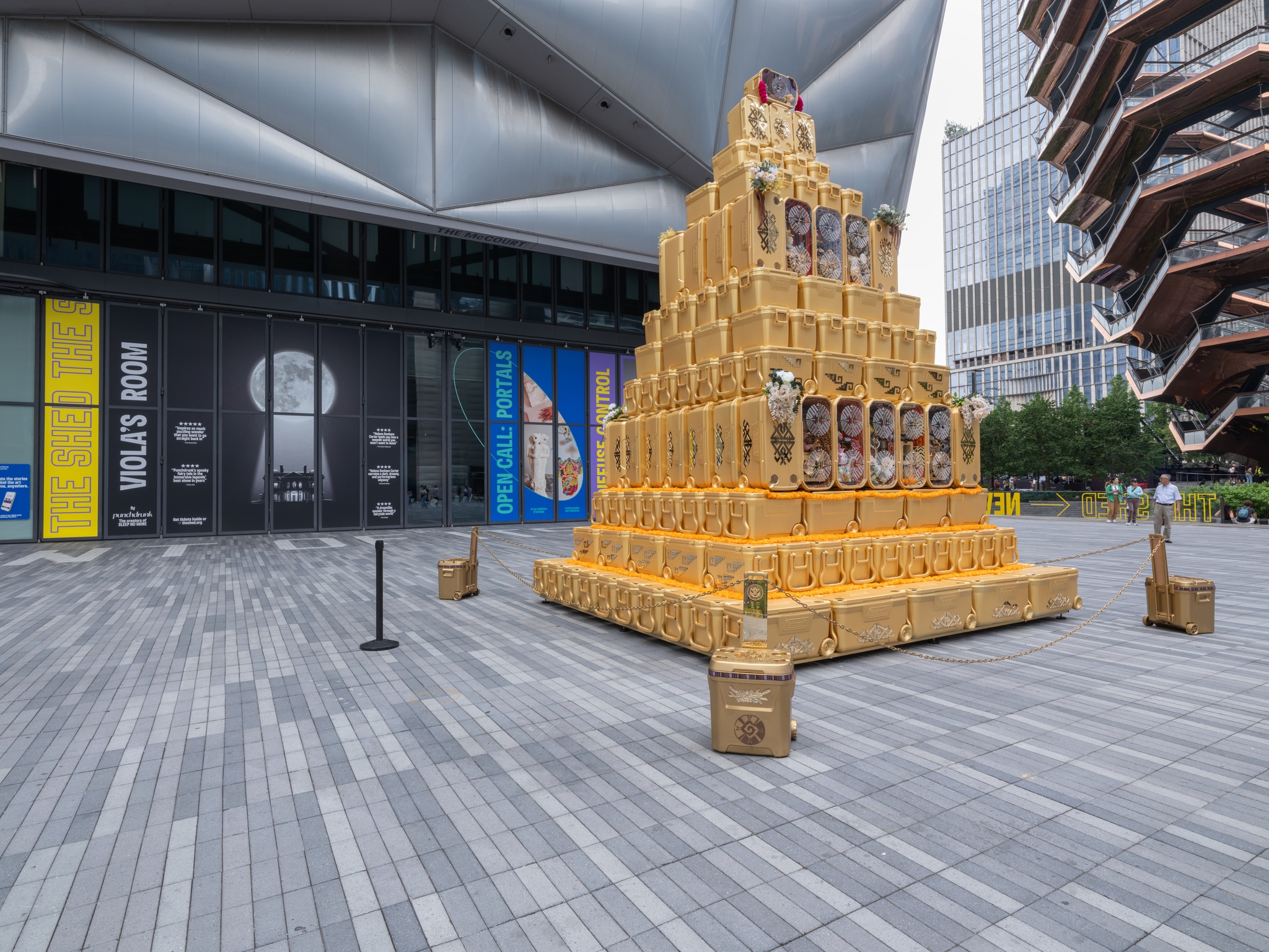 A freestanding, 20-foot-tall golden pyramid glimmers in the sun outdoors on The Shed’s public Plaza. The pyramid is 18 feet square at its base, and seems at first to have a smooth, solid surface. But the pyramid is constructed from 194 gold-painted, matte-plastic rectangular boxes, of different sizes. In fact, these boxes are ice coolers, with their familiar handles, latches and embossed manufacturers’ textures. They are decorated with contrasting stuck-on golden mirrored acrylic glyphs, some protruding more than others.
