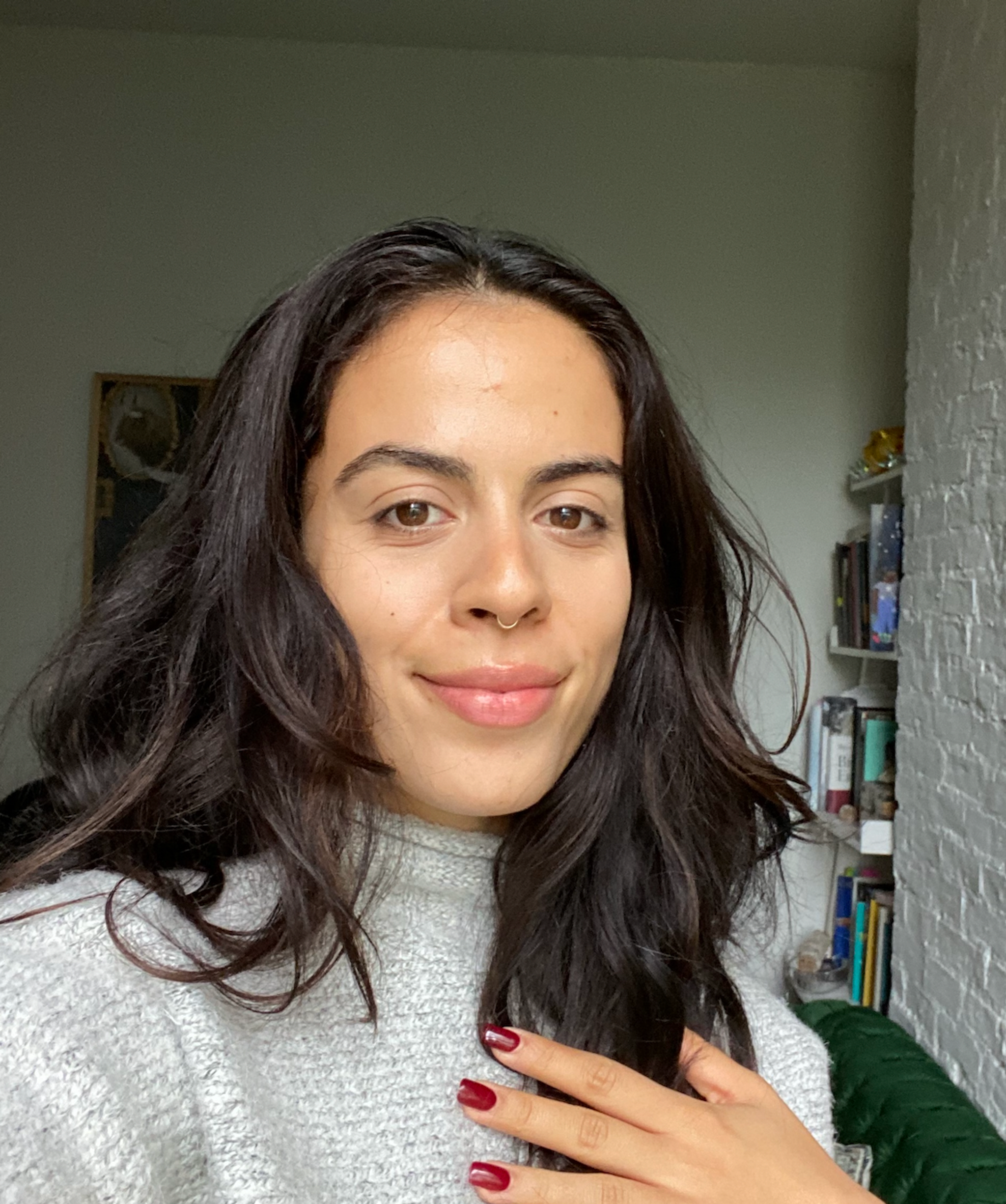 A selfie taken by Maddie Aleman posing in a room with a narrow bookshelf in the corner behind her. She has dark long wavy hair that falls below her shoulders and looks at the camera with one eyebrow raised. She holds one hand to her chest, she wears red fingernail polish. 