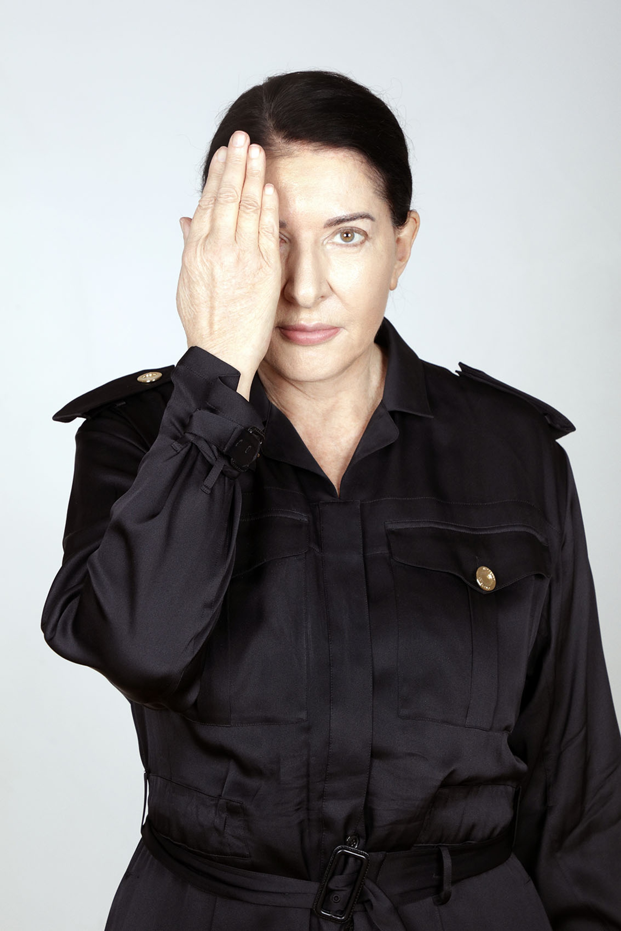 A portrait of artist Marina Abramovic. She has dark hair pulled back behind her head, wears a black trench coat buttoned up, and holds her right hand with the palm covering the right side of her face. 