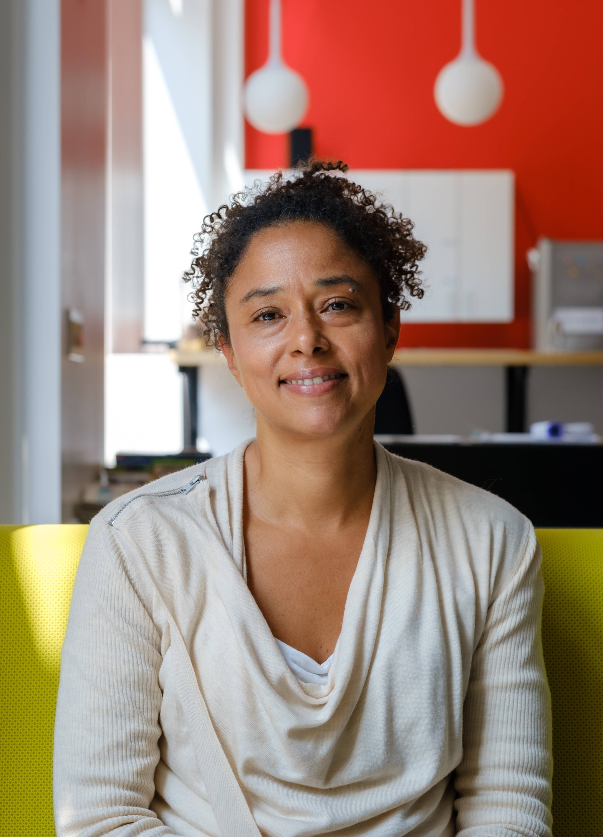 A photo of Deanna Van Buren smiling and seated on a bright yellow sofa. Behind her, pendant lamps hang in the background against a bright red-orange wall. 