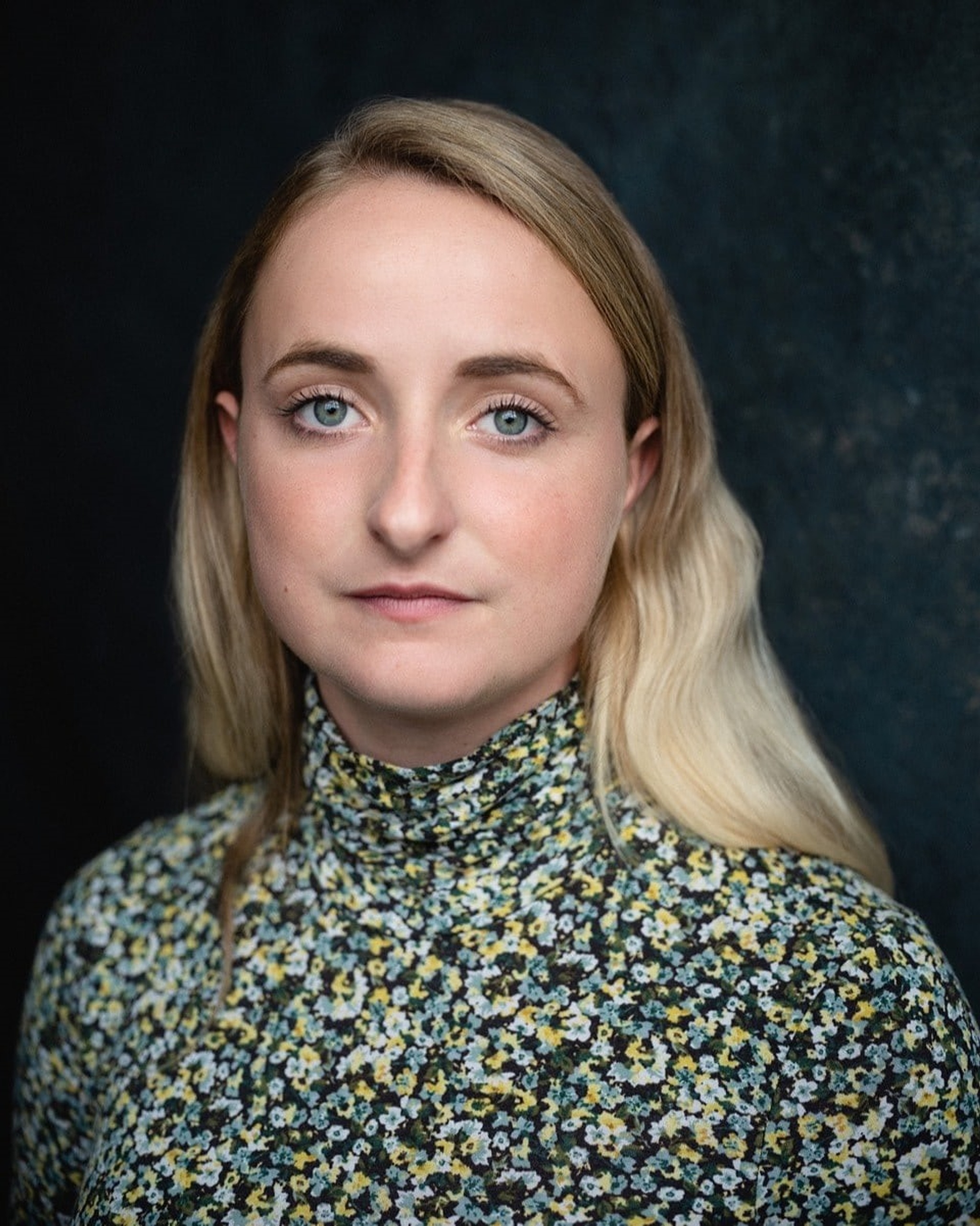 A headshot of actor Eleanor de Rohan, who looks directly at us. Eleanor is white with long blond hair tucked behind her ears. She wears a floral print turtle neck shirt. 