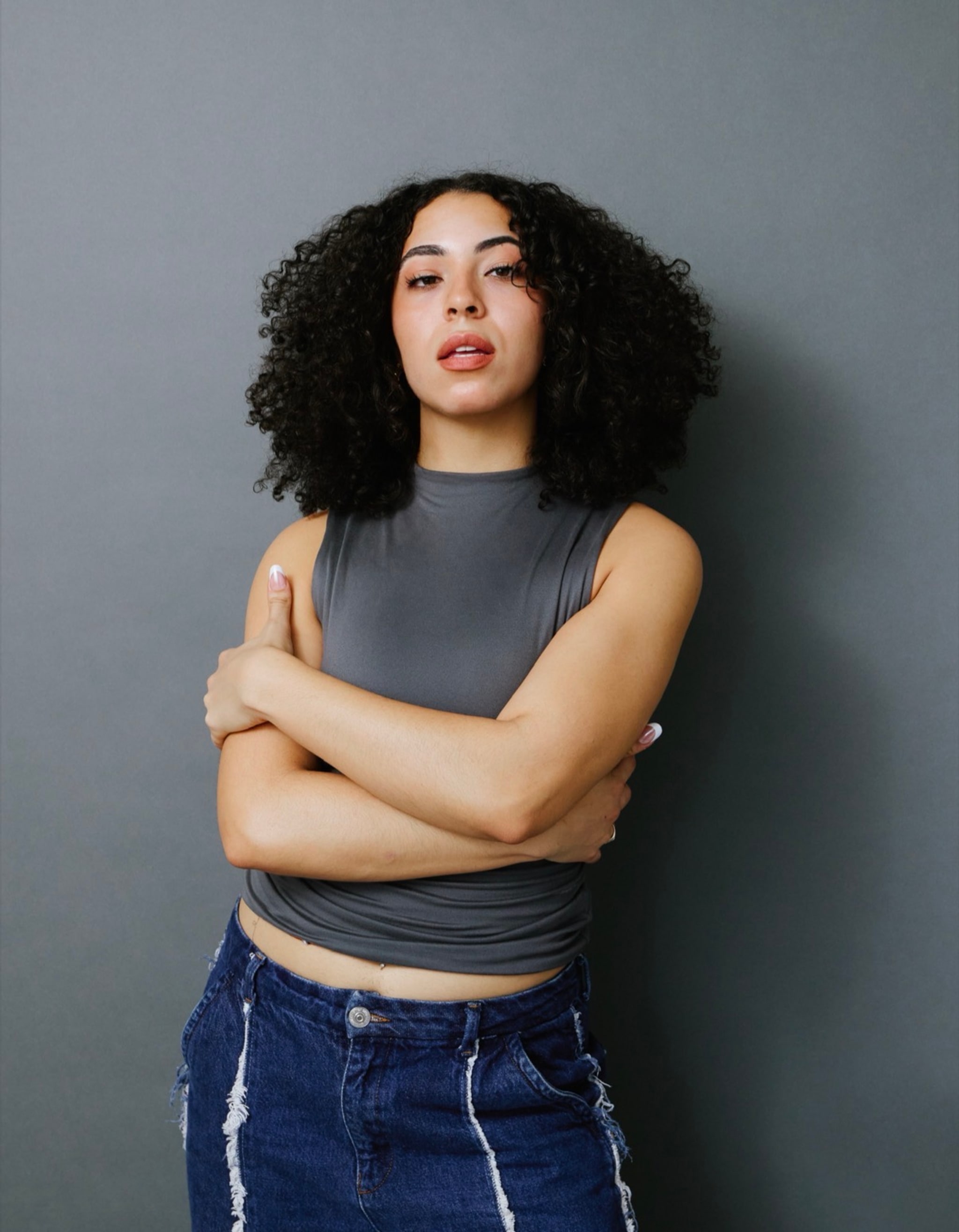 A portrait of performer Ava Jones who stands against a gray wall with arms crossed and head tilted back slightly while looking at us. Ava has curly dark brown hair parted down the middle and wears a gray sleeveless shirt and blue jeans. 