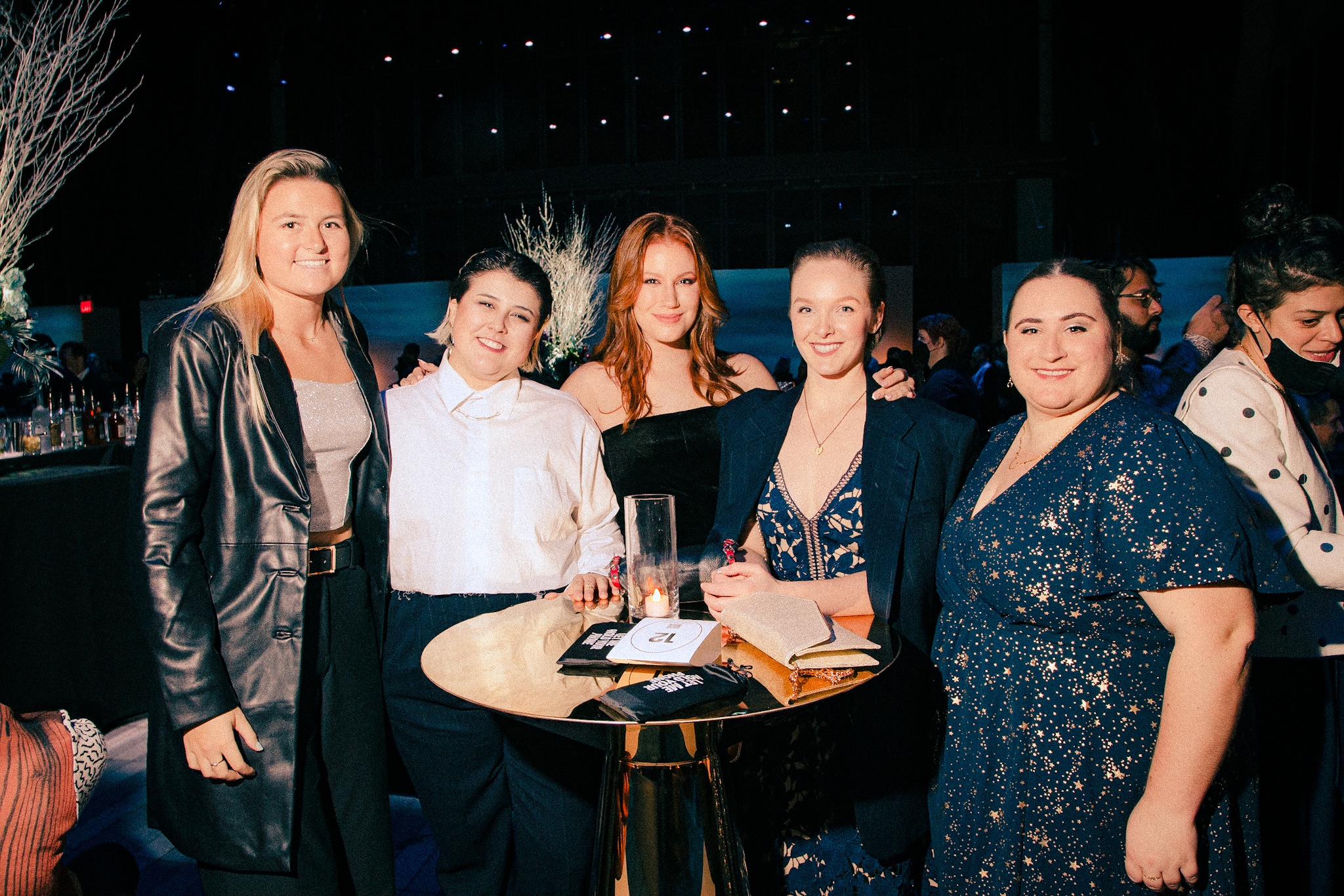 A group of guests at The Shed's 2021 Gala stand with their arms around each other around a high top table. 