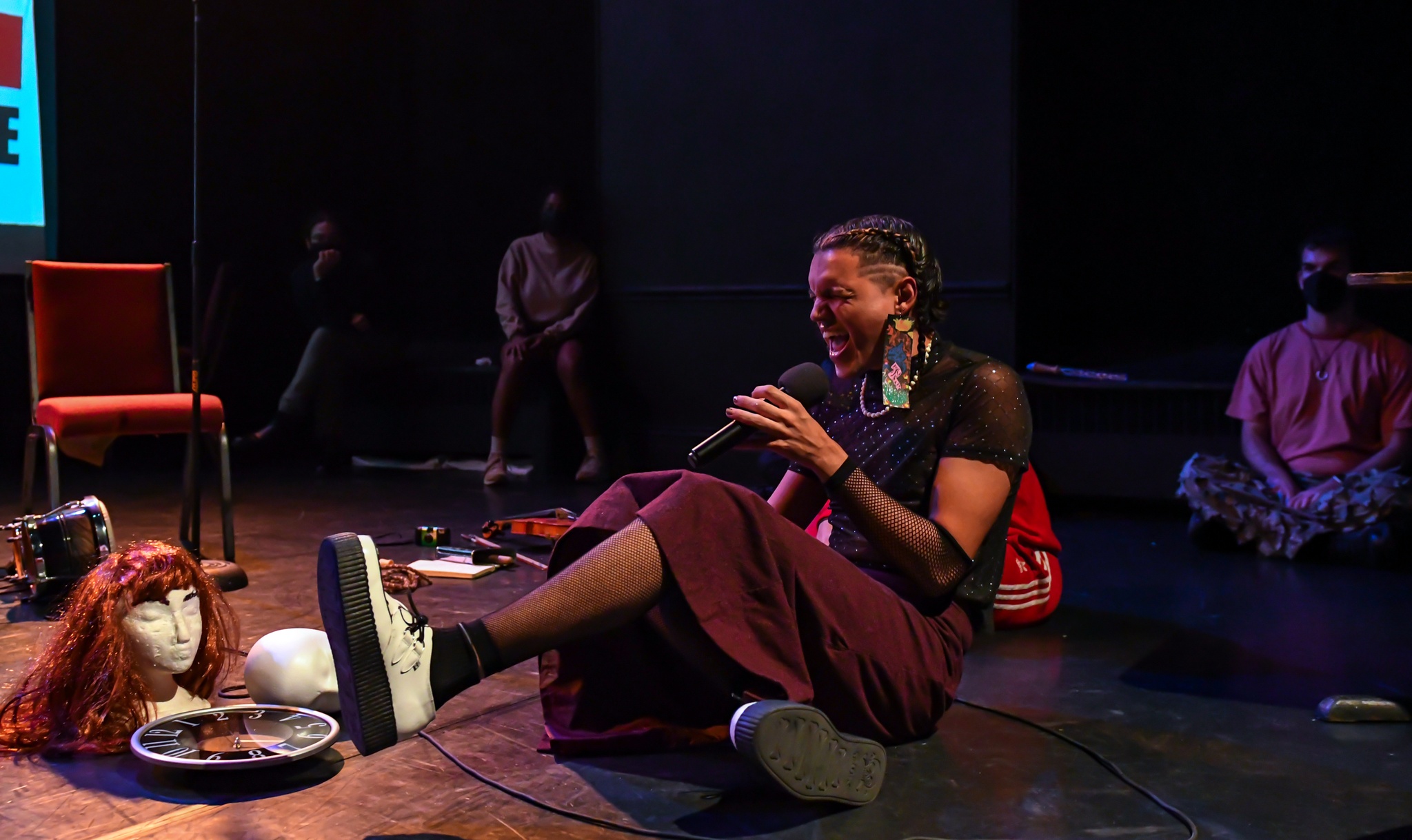 Mobéy Lola Irizarry sits on the ground singing passionately into a microphone. They are a genderqueer Puerto Rican and Chickasaw person who wears a long skirt, platform shoes, with their dark brown hair in braids running tightly down the sides of their head. 