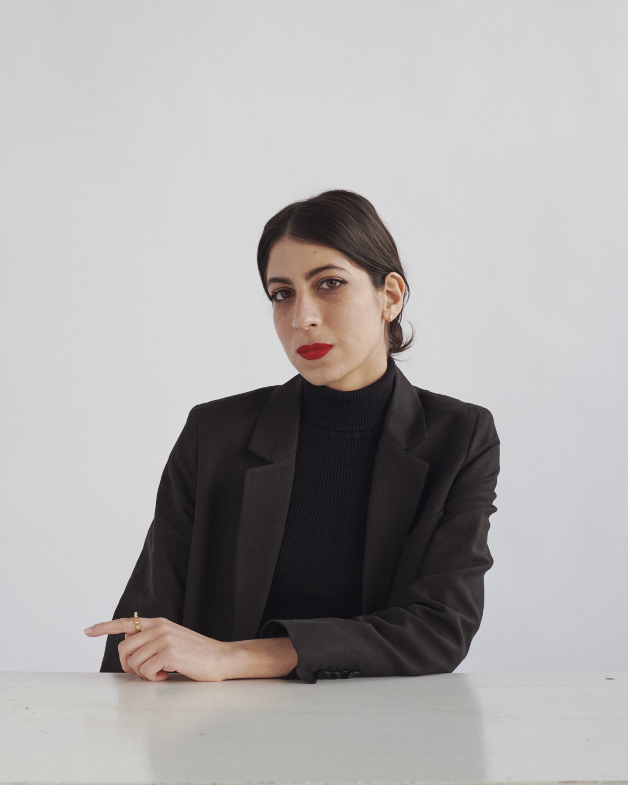 A portrait of Natalia Viera Salgado, a Puerto Rican woman who poses at a white table against a white backdrop. She leans one arm on the table and looks directly at us. She has short dark brown hair tucked behind her ears.