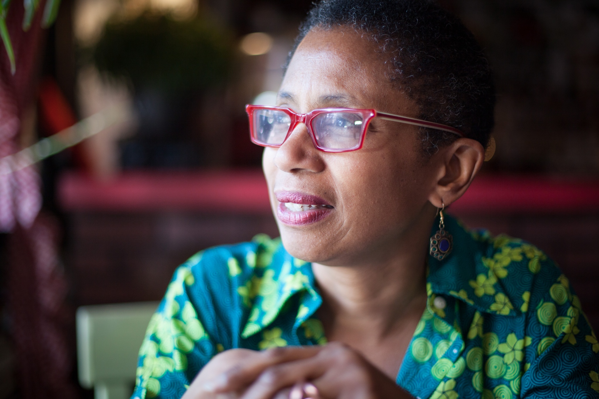 A photo of Walis Johnson wearing red eyeglasses and looking out of the frame of the image. She wears a green shirt and light falls on her face as if from a window. 