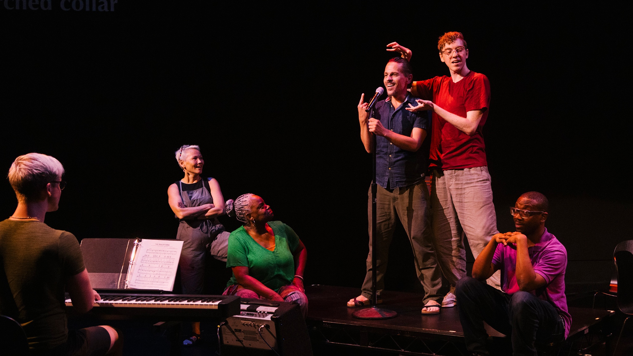 Six Deaf performers appear on stage in Garrett Zuercher's "Inside/Look." One central performer stands at a microphone while the others look toward him. In the foreground, one of the performers is seated playing a piano with their back to us. 