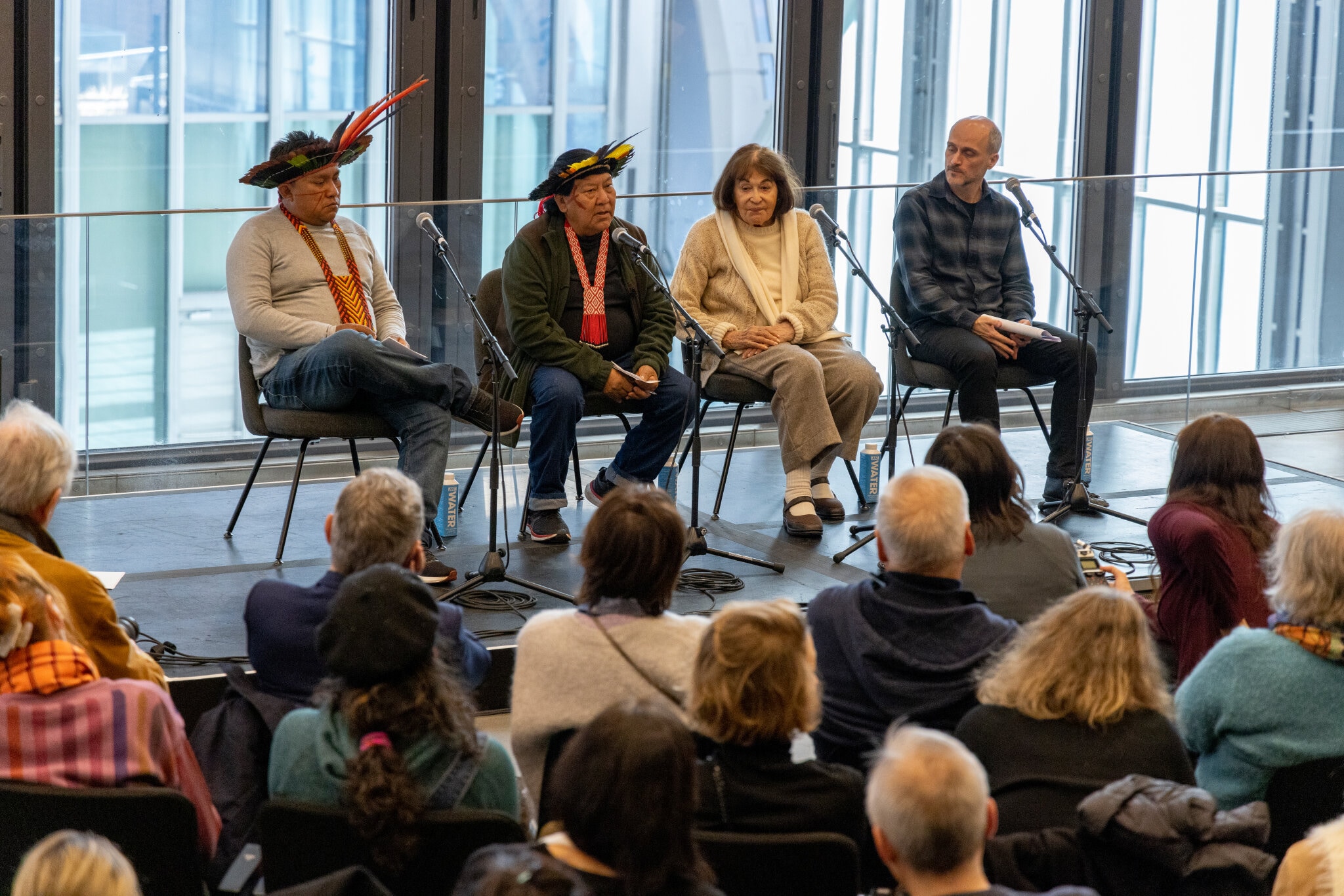 Four panelists sit on a stage with large windows behind them. On the left are Dario and Davi Kopenawa, two Yanomami men who wear head dresses made of colorful feathers. On the right of them sits artists Claudia Andujar, a Brazilian white woman with brown hair wearing an off white outfit. To her right is curator Thyago Nogueira, a white Brazilian man wearing a dark button down shirt in a check plaid pattern.