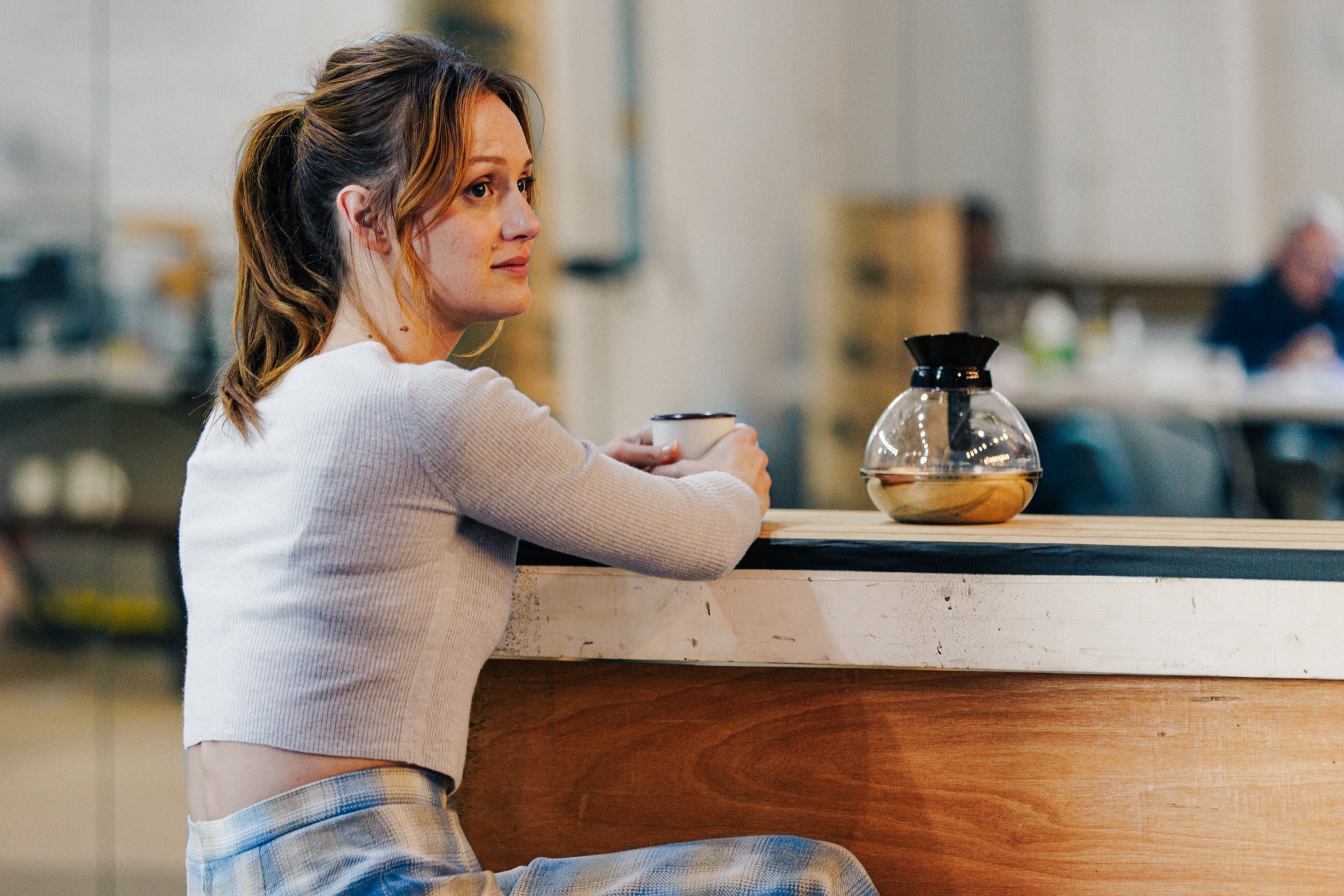 Actor Kerry Bishé, a white woman with light brown hair in a pony tail, sits at a counter in a rehearsal space. She looks to the side, seen from behind, and an almost empty coffee pot sits on the counter in front of her. 