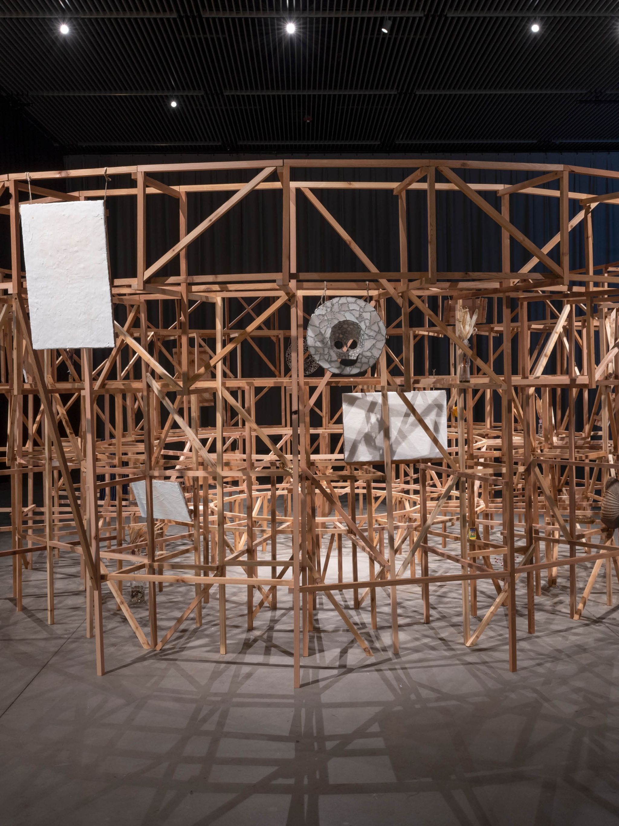 A wooden sculptural installation in a gallery with low lighting. The circular structure, about 6 feet high, is a scaled down model of the architectural support for arena seating. It is made of thin beams of wood and has objects like masks and paintings hanging from it facing outward. The center of the arena is empty and inaccessible to gallery visitors. 