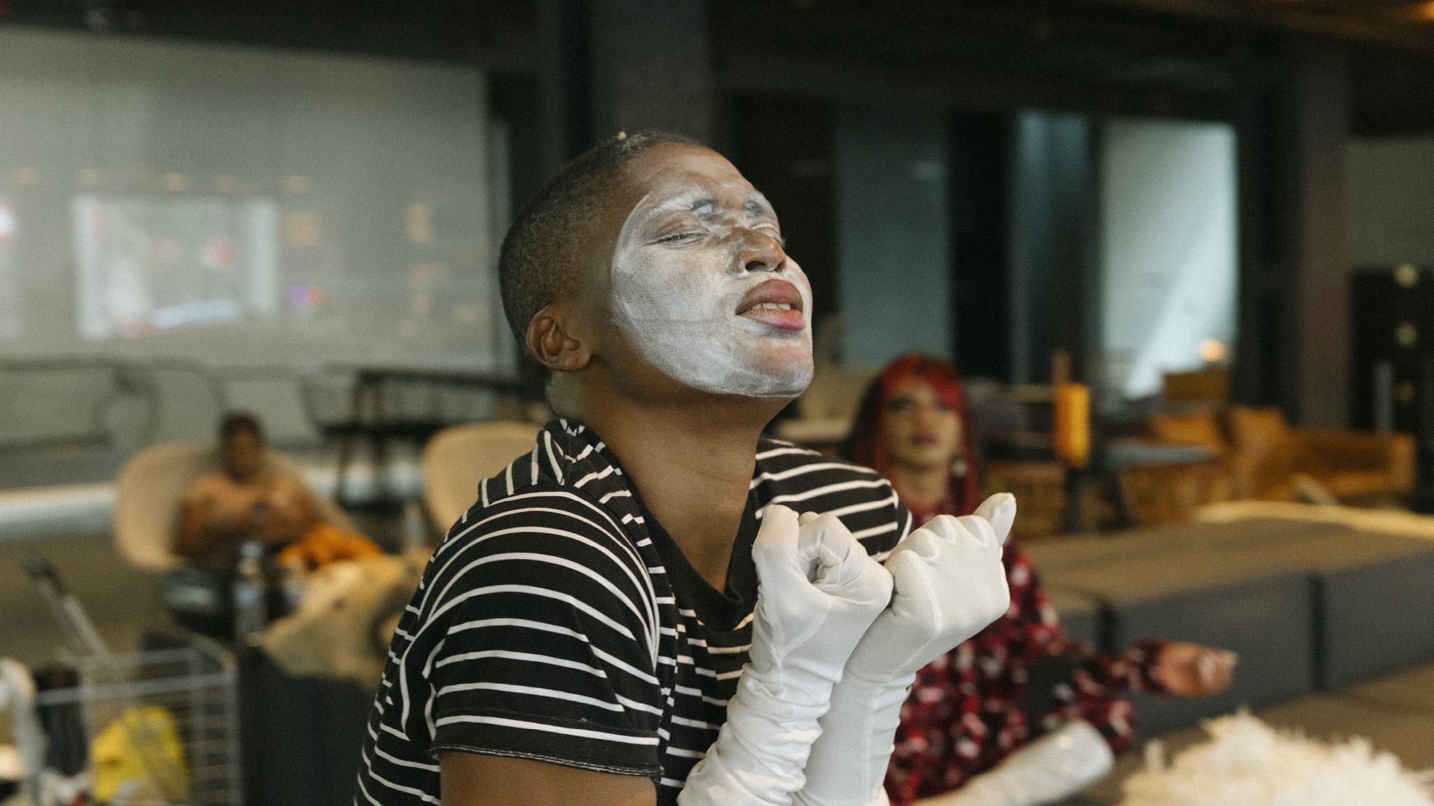 NIC Kay poses in character with white mime makeup on their face. They are in The Shed's lobby with couches and low seats behind them. They hold their hands up to their chest, wearing long white gloves, and pucker their face, looking upward. 