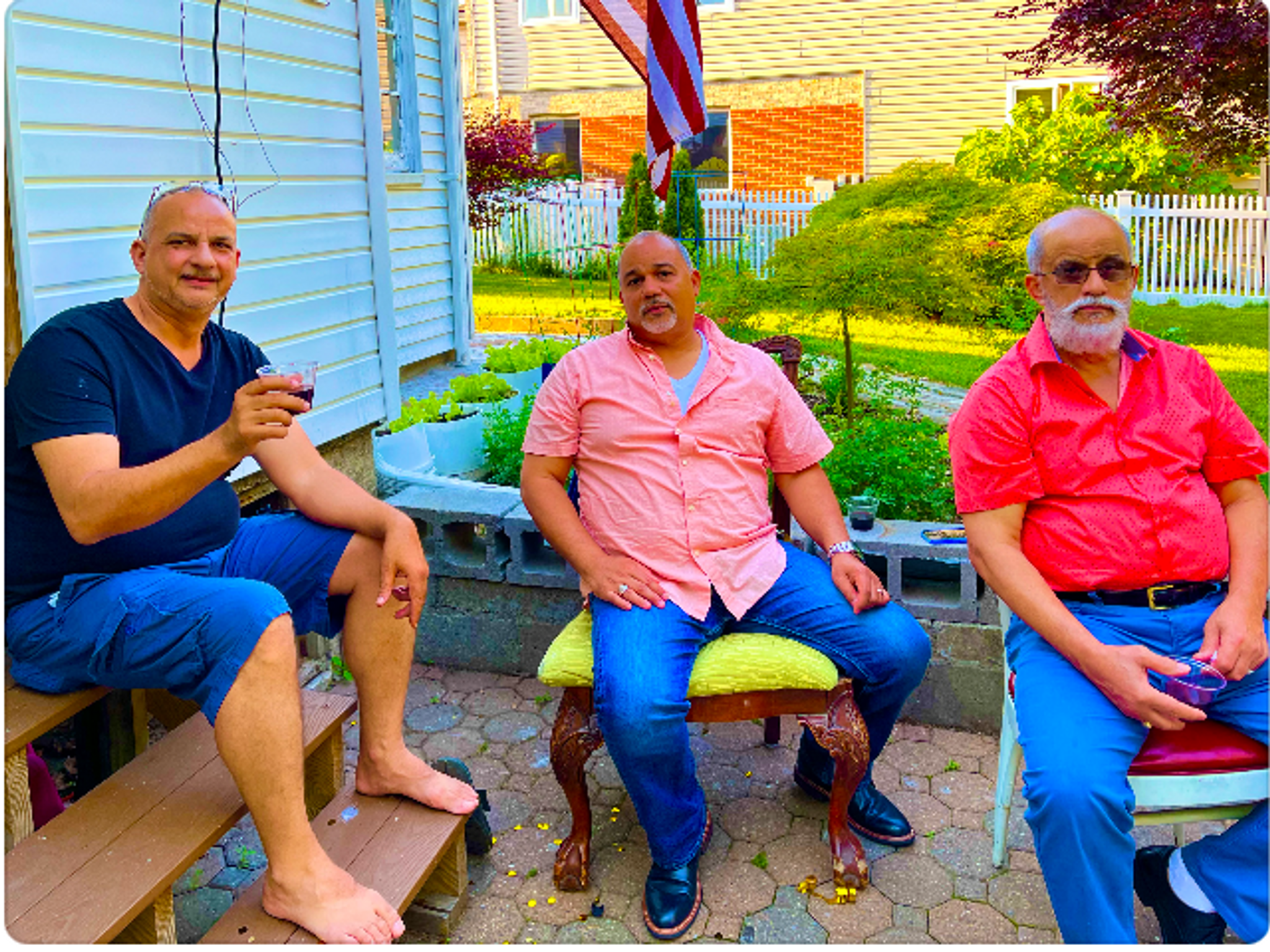Three middle aged Dominican men sit in a suburban backyard. They are seated in a row, with one of them resting on steps leading up to a house, one on a dining room chair with a lime green upholstered seat, and the third on a metal chair. The men wear a blue tshirt, pink button down shirt, and bright red button down shirt. Behind them is a green lawn.