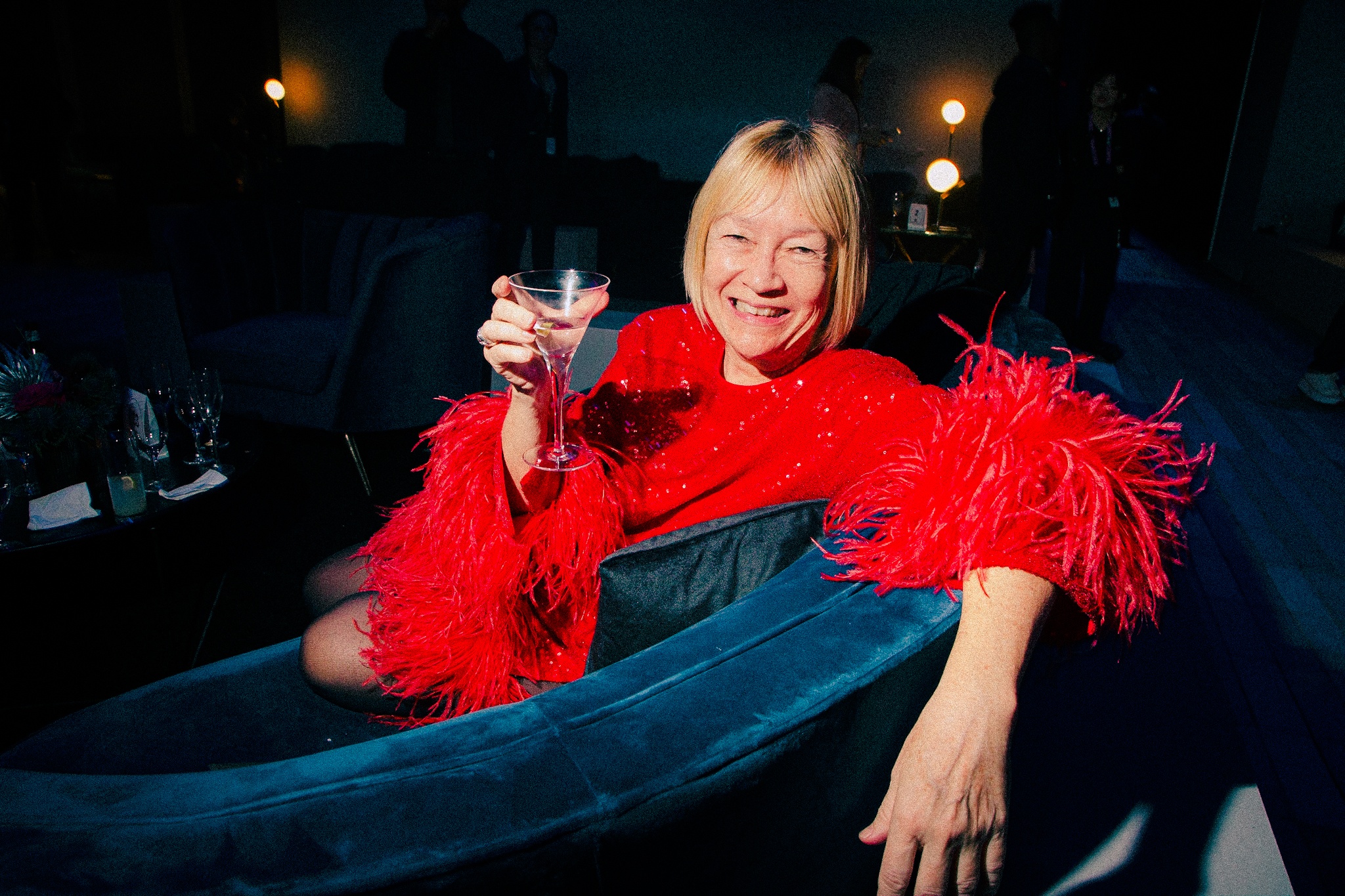 A gala guest reclines on a plush sofa in a lounge, raising her glass as if giving a toast.