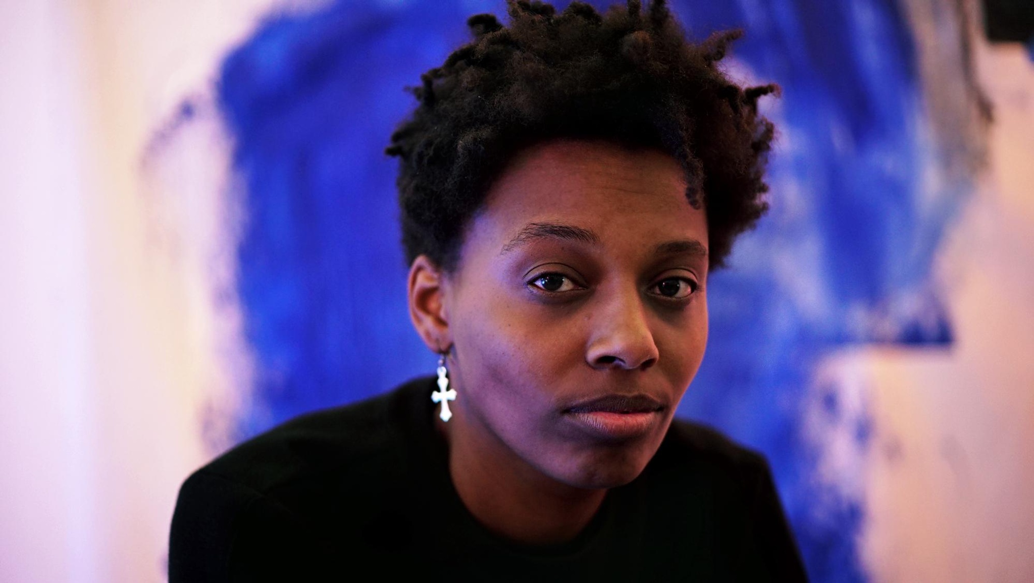 A photo of the artist Le'Andra LeSeur in front of a blue painting blurry in the background. LeSeur has short hair and wears a cross earring.