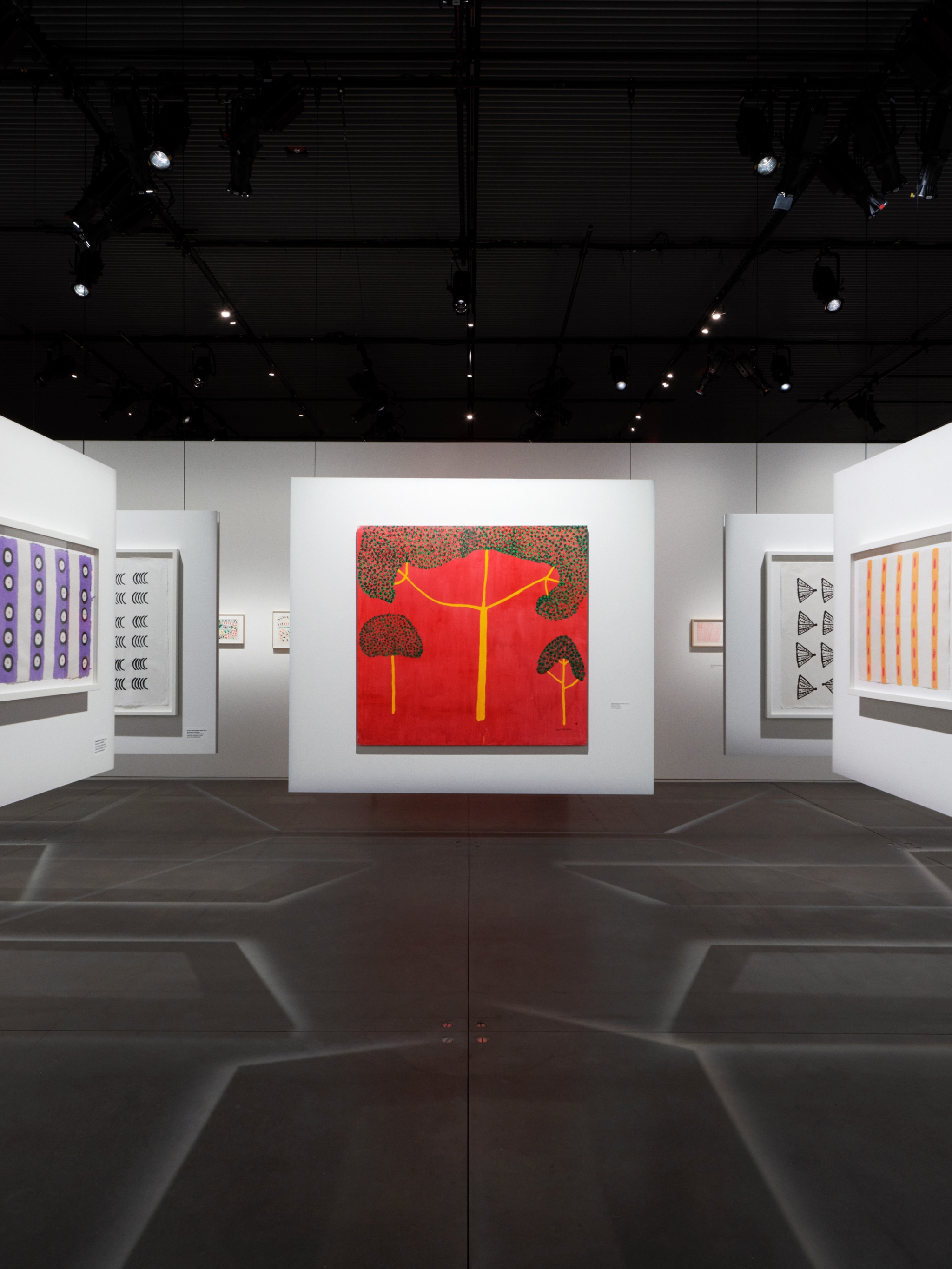 An art gallery with drawings and paintings on walls suspended from the ceiling and on fixed walls. The suspended walls make shadows of intersecting lines on the gray concrete floors. Situated prominently in the center of the gallery is a painting of three trees with yellow trunks and green canopies against a bright red background.