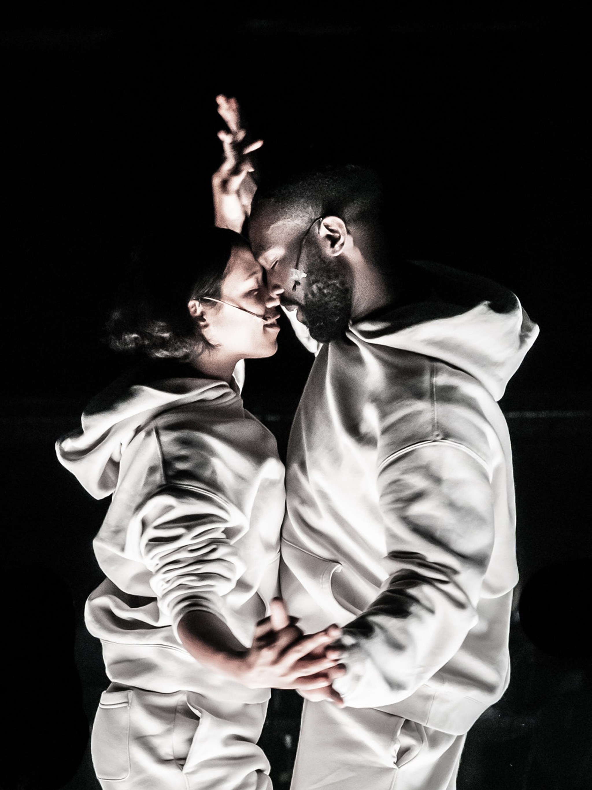 The two actors Taylor Russell and Paapa Essiedu face each other pressing their foreheads together. We see them from the shoulders up in a close up. They wear gray hoodies and clasp hands raised behind them. Their eyes are closed and they both have slight smiles on their faces.