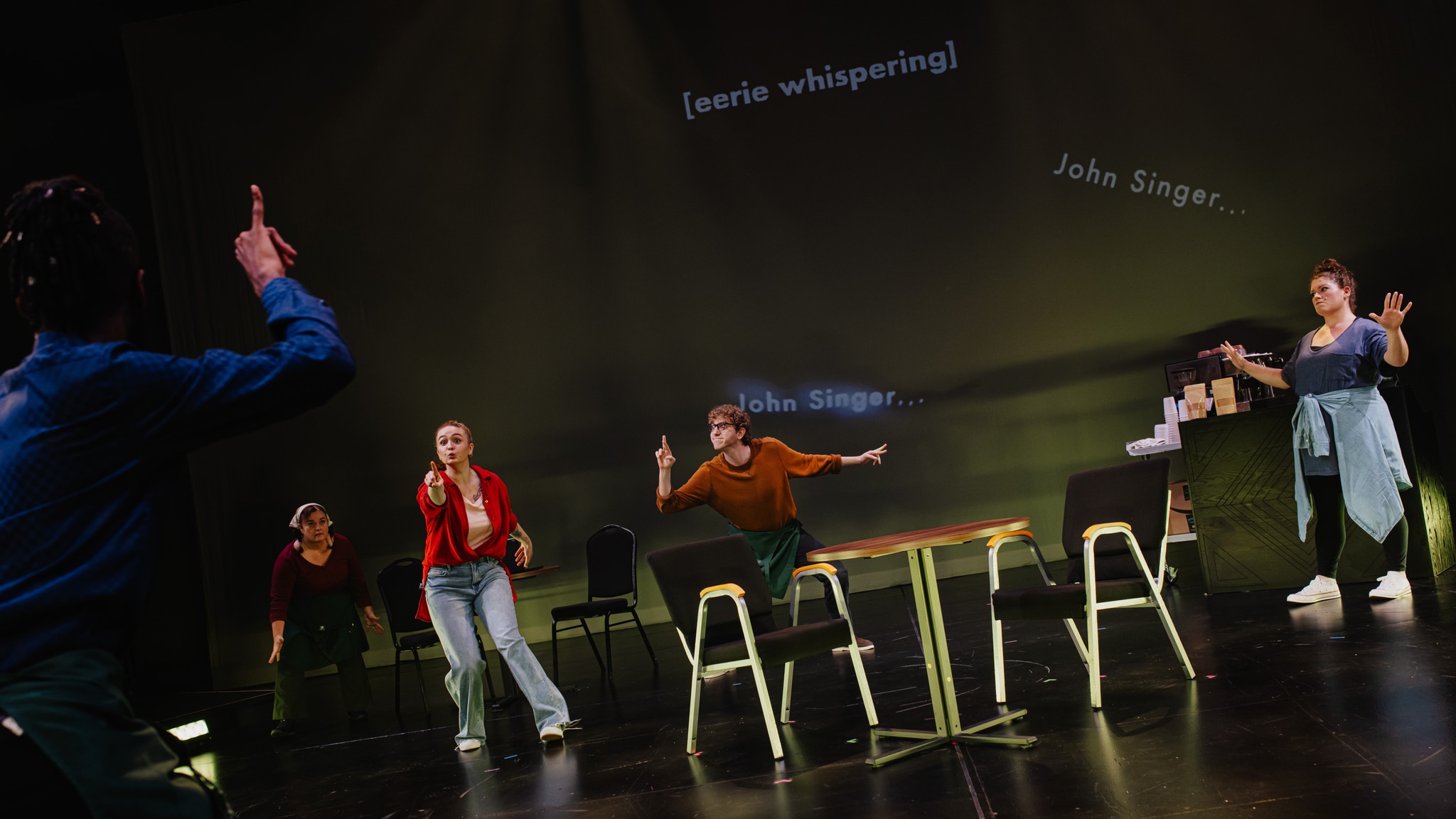 Deaf actors stand spread across a dark stage with tables and chairs representing a coffee shop. On a screen behind them are open captions reading [eery whispering] and John Singer.
