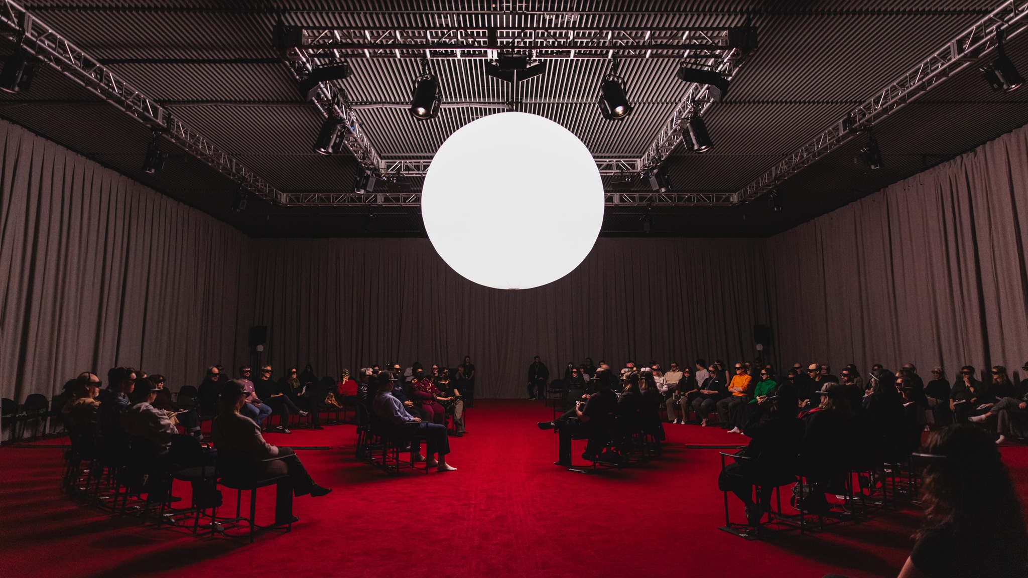A large curtained performance space with a bright red carpet. Audience sit in concentric circles wearing mixed reality headsets. At the center of the room hangs a glowing white orb-like light fixture. 