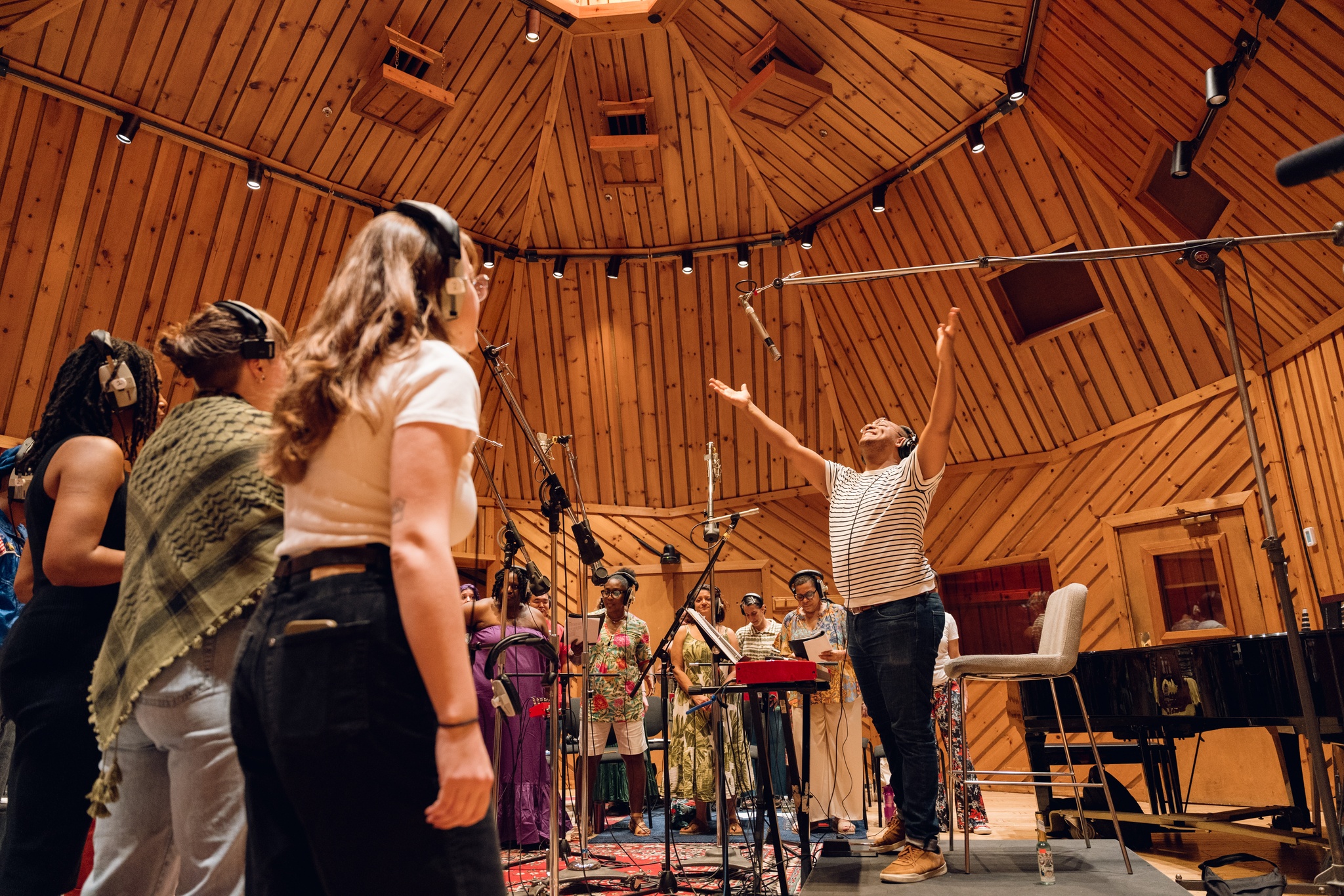 Troy Anthony stands in front of choir members with his arms both spread out joyously above his head. The choir members are seen from behind, with their attention on Troy. 