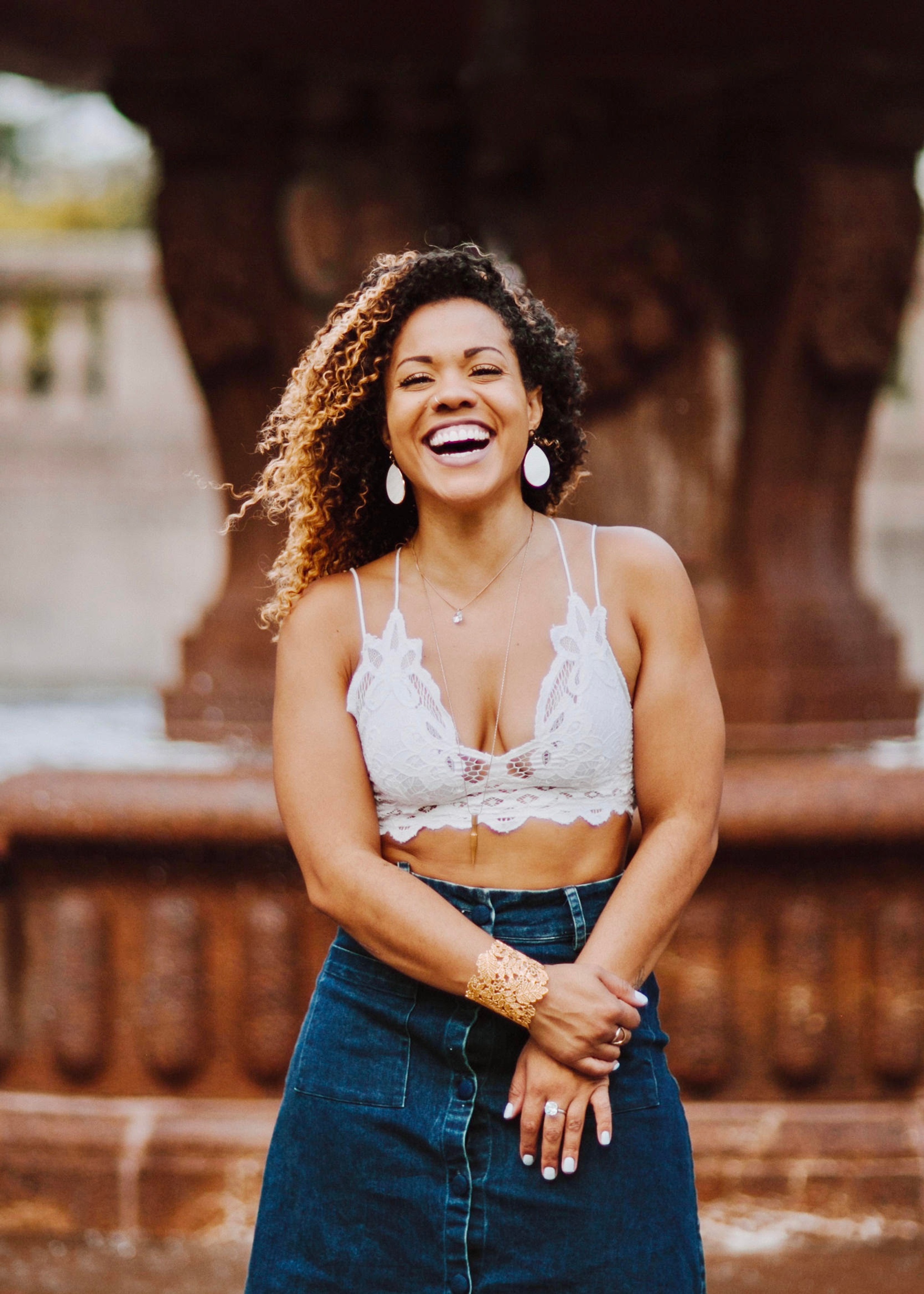 A woman with curly brown hair slightly blown back in the wind while she holds her hands in front of her waist. She wears a white crop top and jean skirt. 