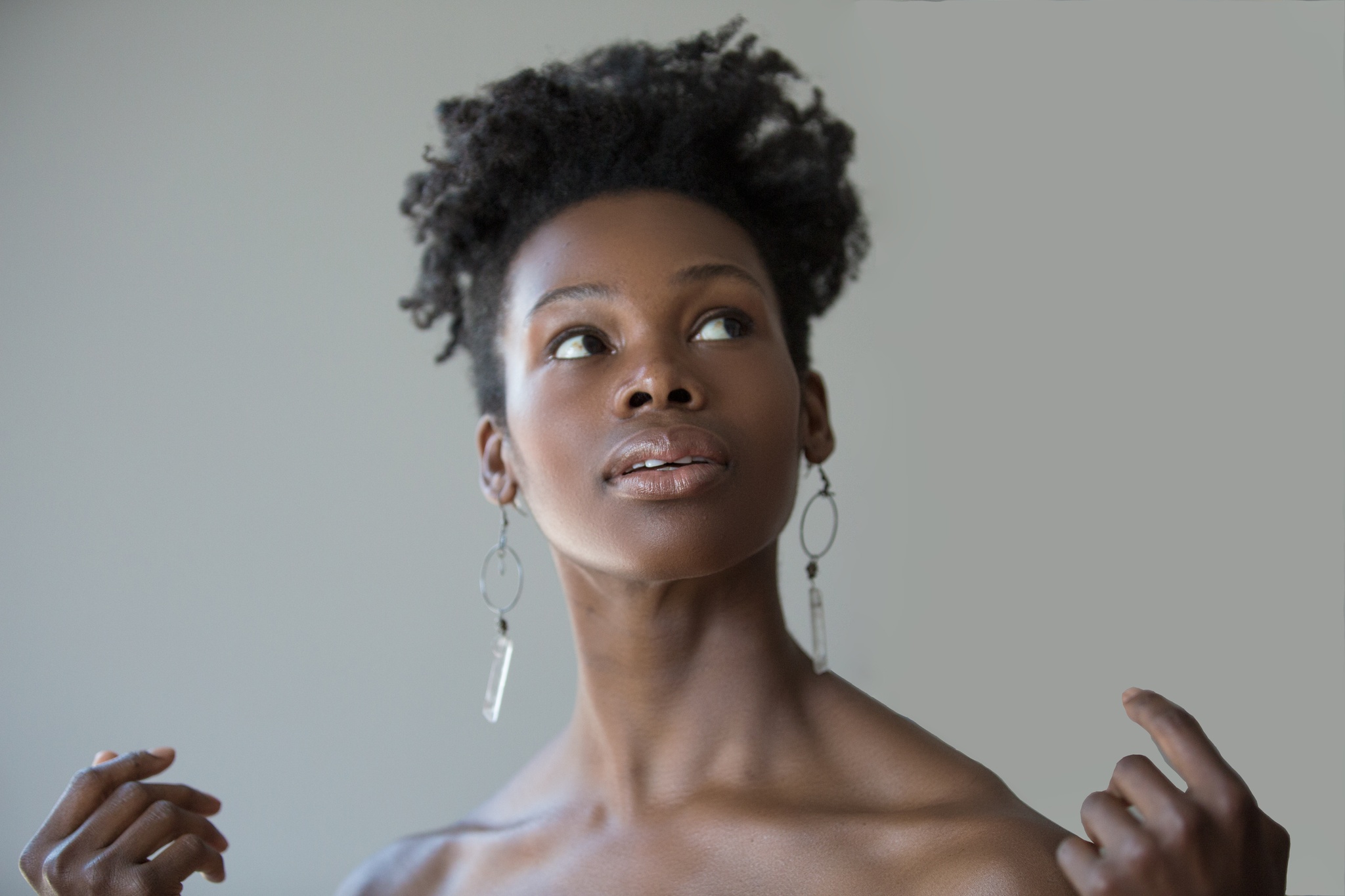 A Black woman pictured from the shoulders up. Her shoulders are bare and her hands are raised to just appear above the bottom frame of the image. She looks up at something above her. 