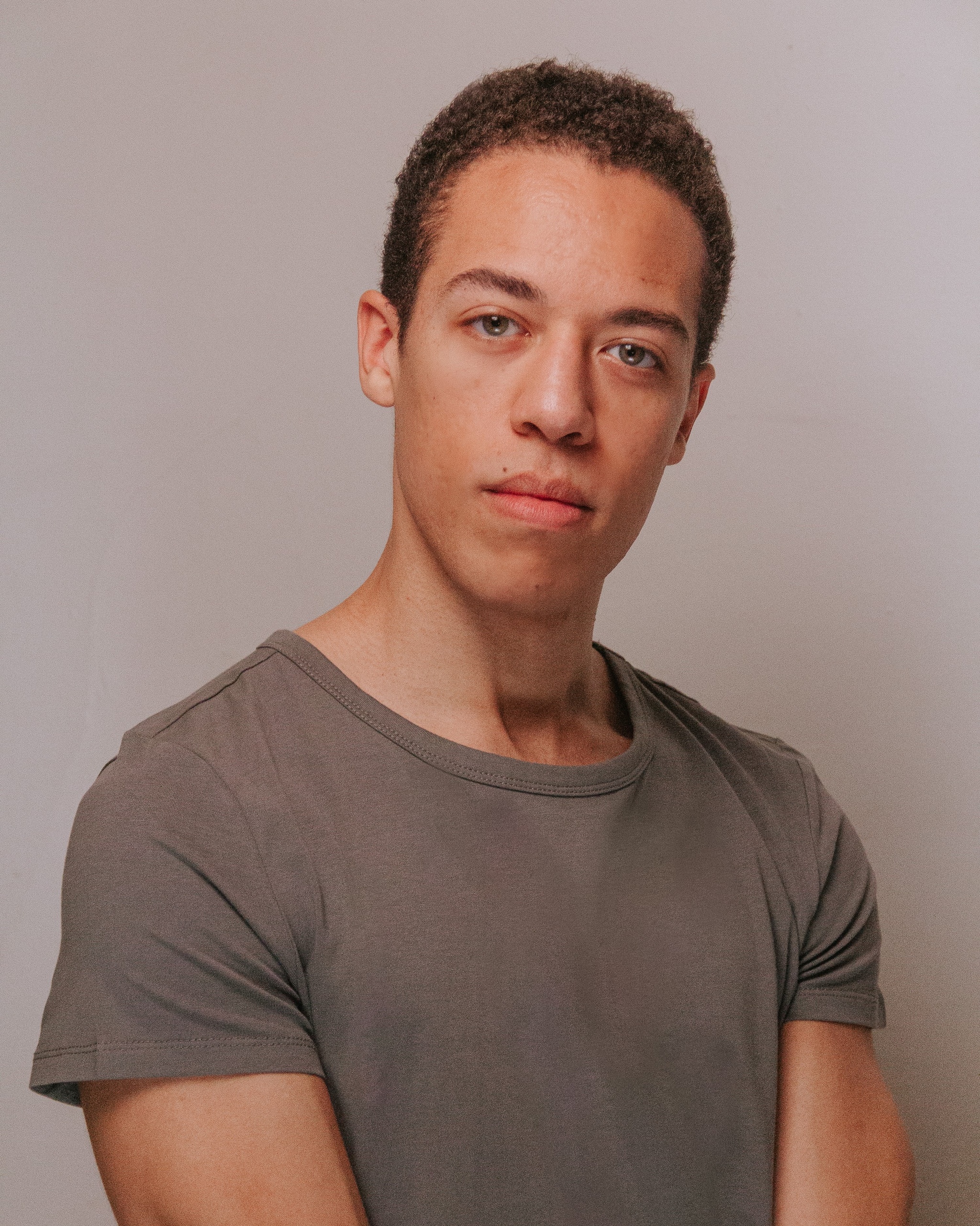 A person with short brown hair seen from the chest up, with arms at his sides and his head at a slight angle while he looks at the camera. He stands against a neutral background. 