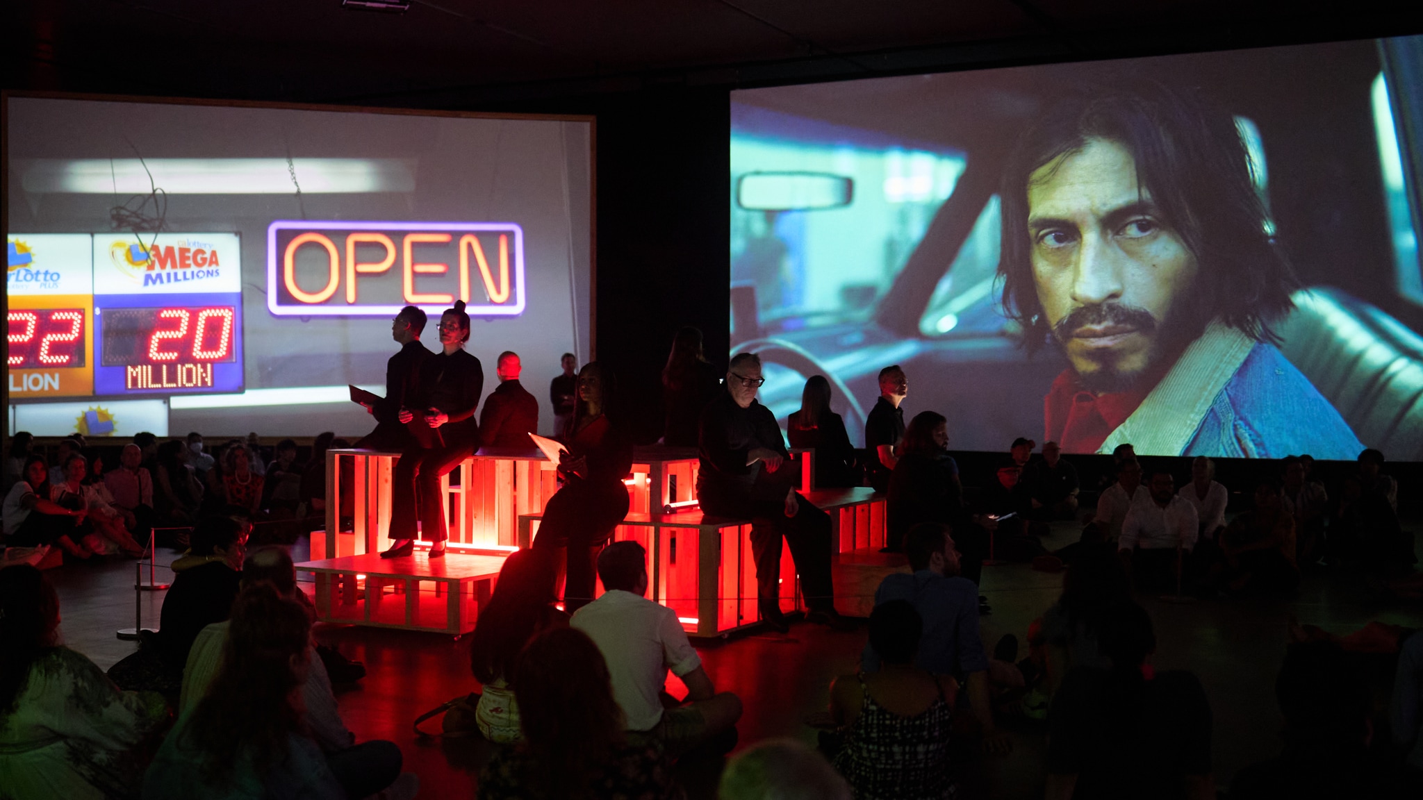 A group of eight chorus members dressed in black sit on a platform at the center of a dark gallery with large film screens behind them. The platform is illuminated by red light and the film screens show images of a convenience store's neon open sign and a man peering out of the driver's window of a pickup truck. 