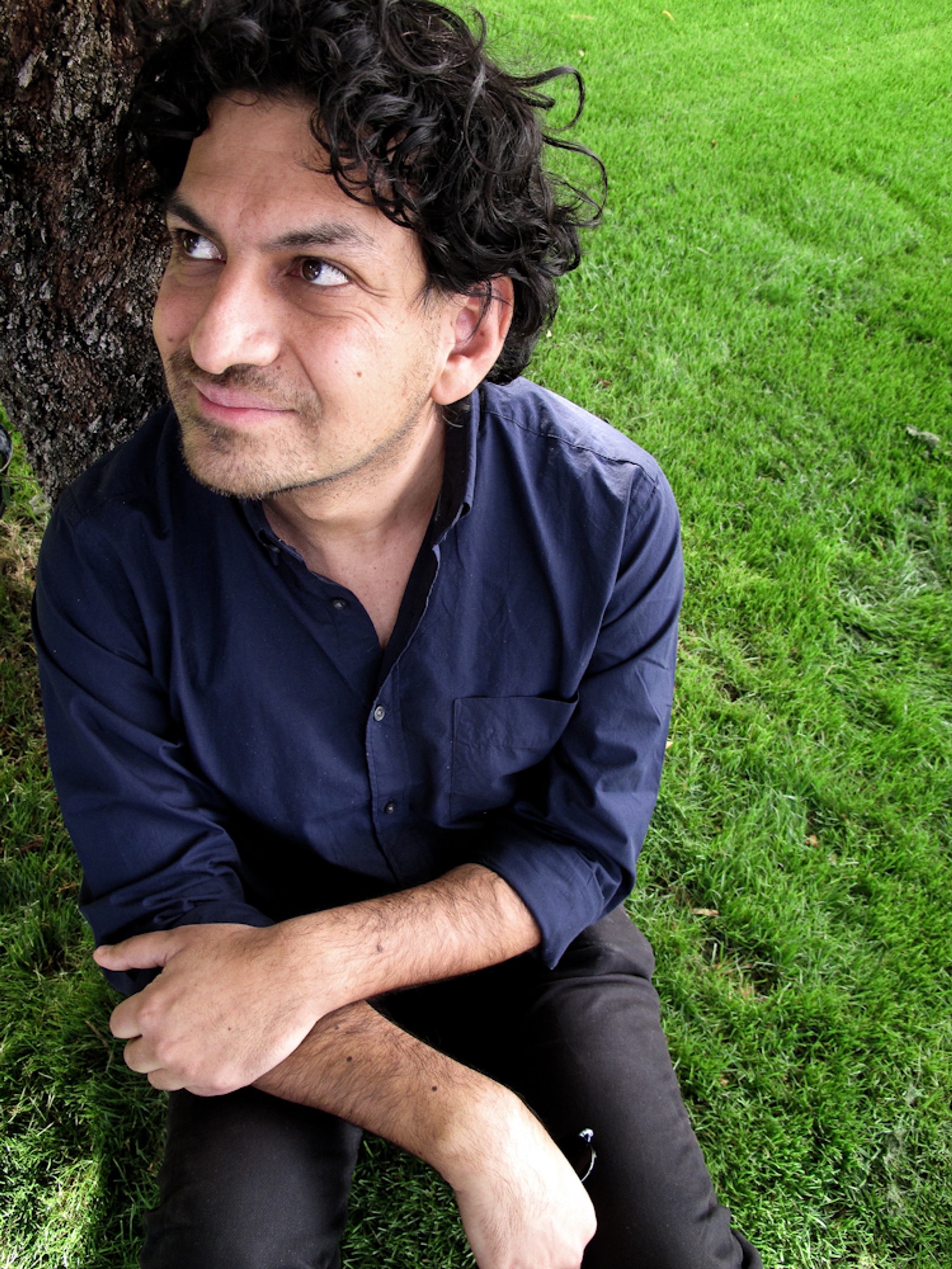 A photo of Carlos A. Gutierrez, taken from above. Gutierrez sits on green grass against the trunk of a tree. 