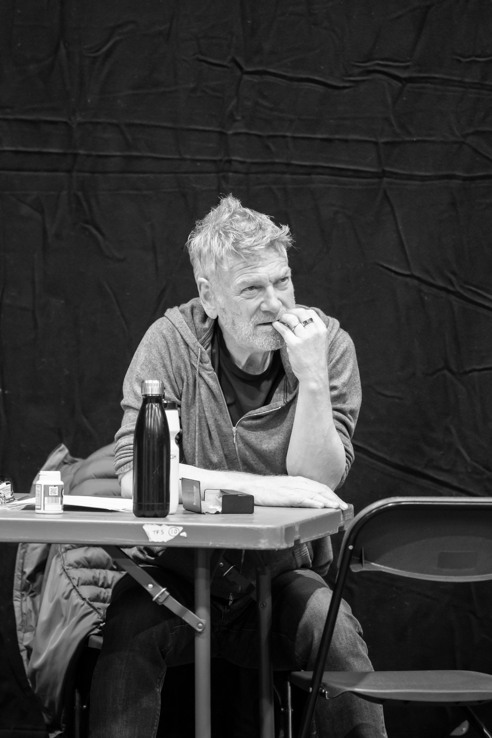 Kenneth Branagh sits in rehearsal at a table. He gives his attention to something happening out of frame, with one hand held up to his chin.