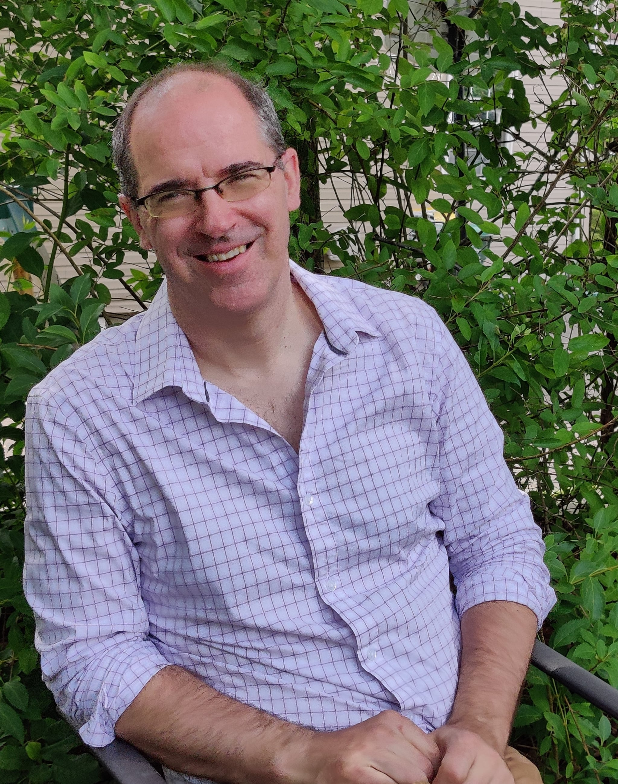 A white man in a lilac shirt leans to the side and smiles in front of green trees and bushes.
