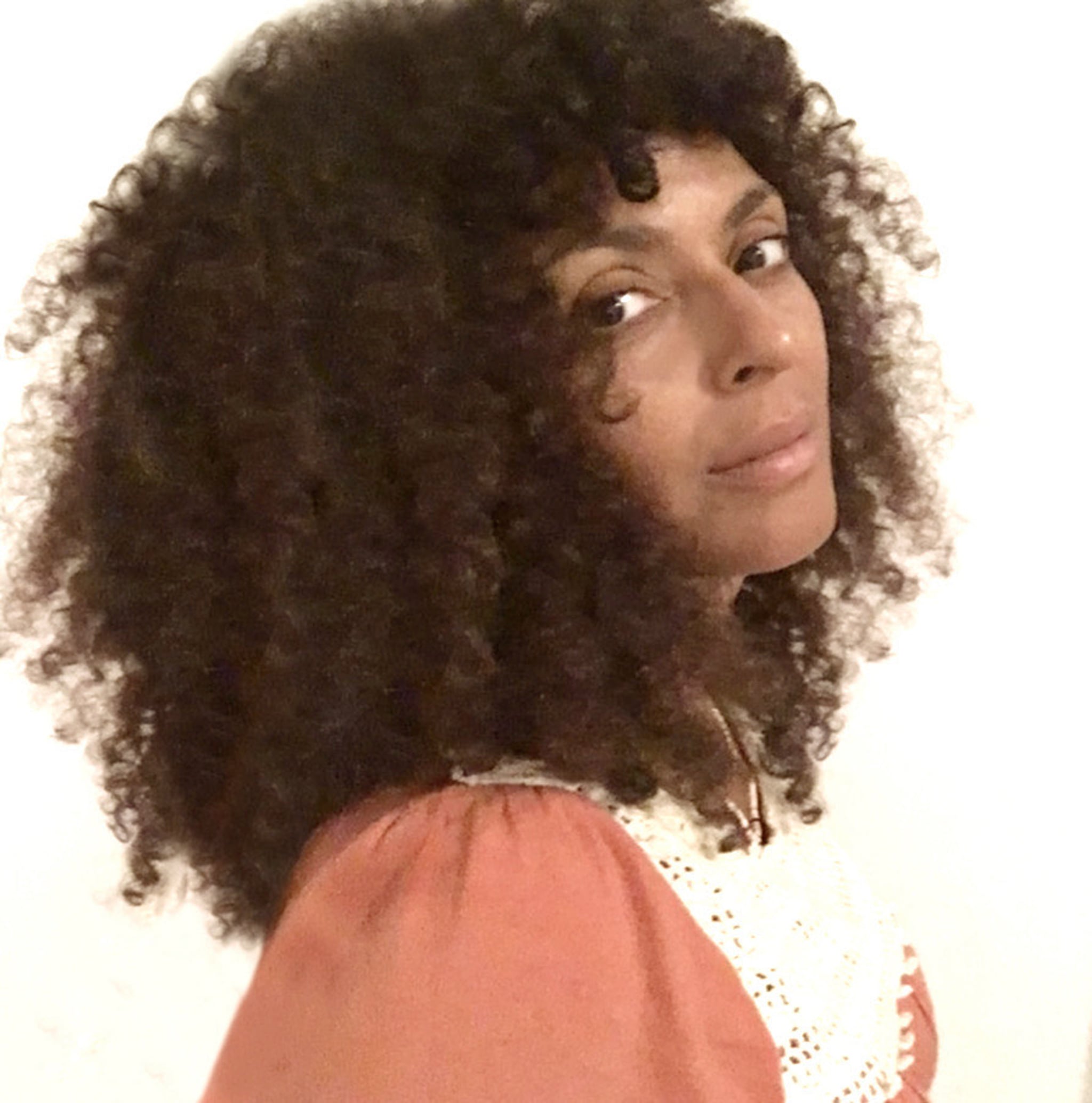 A portrait of DJ Kamala looking over her shoulder. She has curly shoulder-length brown hair and wears a peach-colored shirt. 