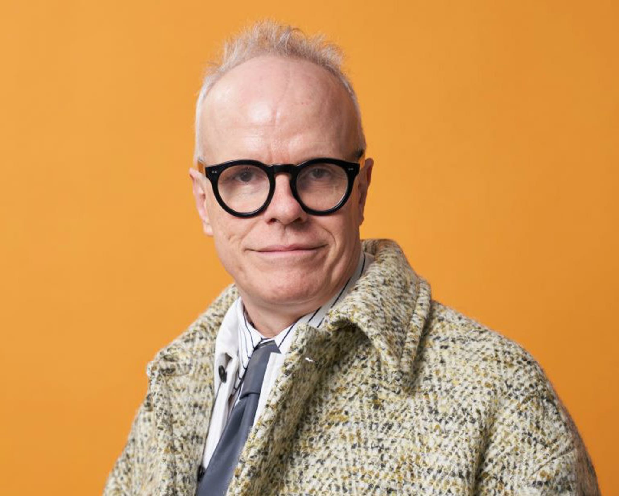 Curator Hans Ulrich Obrist, a white man with thin gray hair, poses for a portrait in front of a deep yellow background. He looks directly at us through black, thick-rimmed glasses and smiles slightly. 