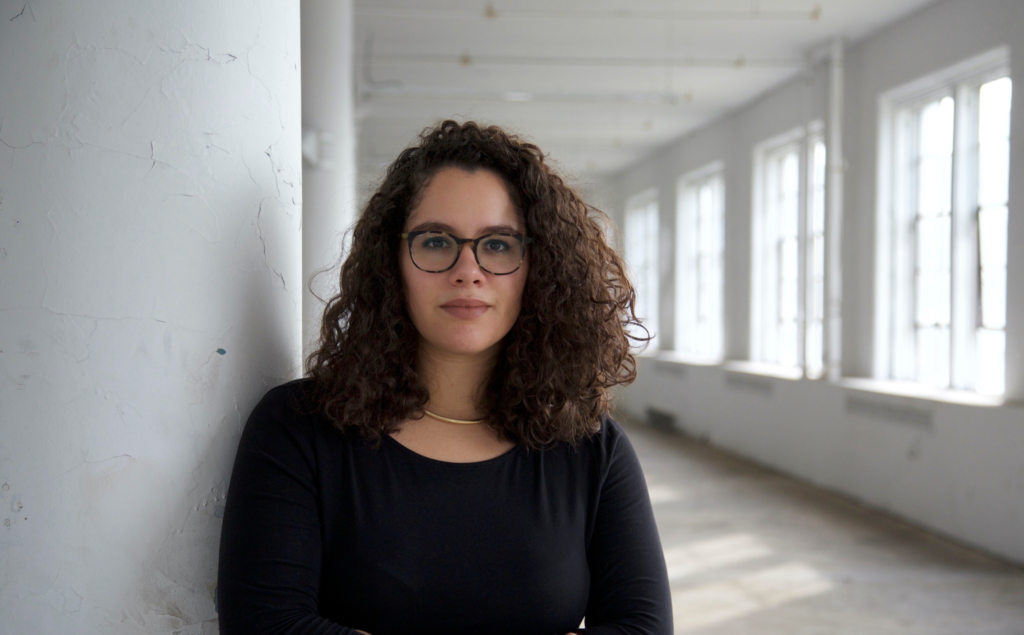 A photo of Alex Santana, who has shoulder-length hair, wears glasses, and poses in a hallway with sunny windows receding into the background. 