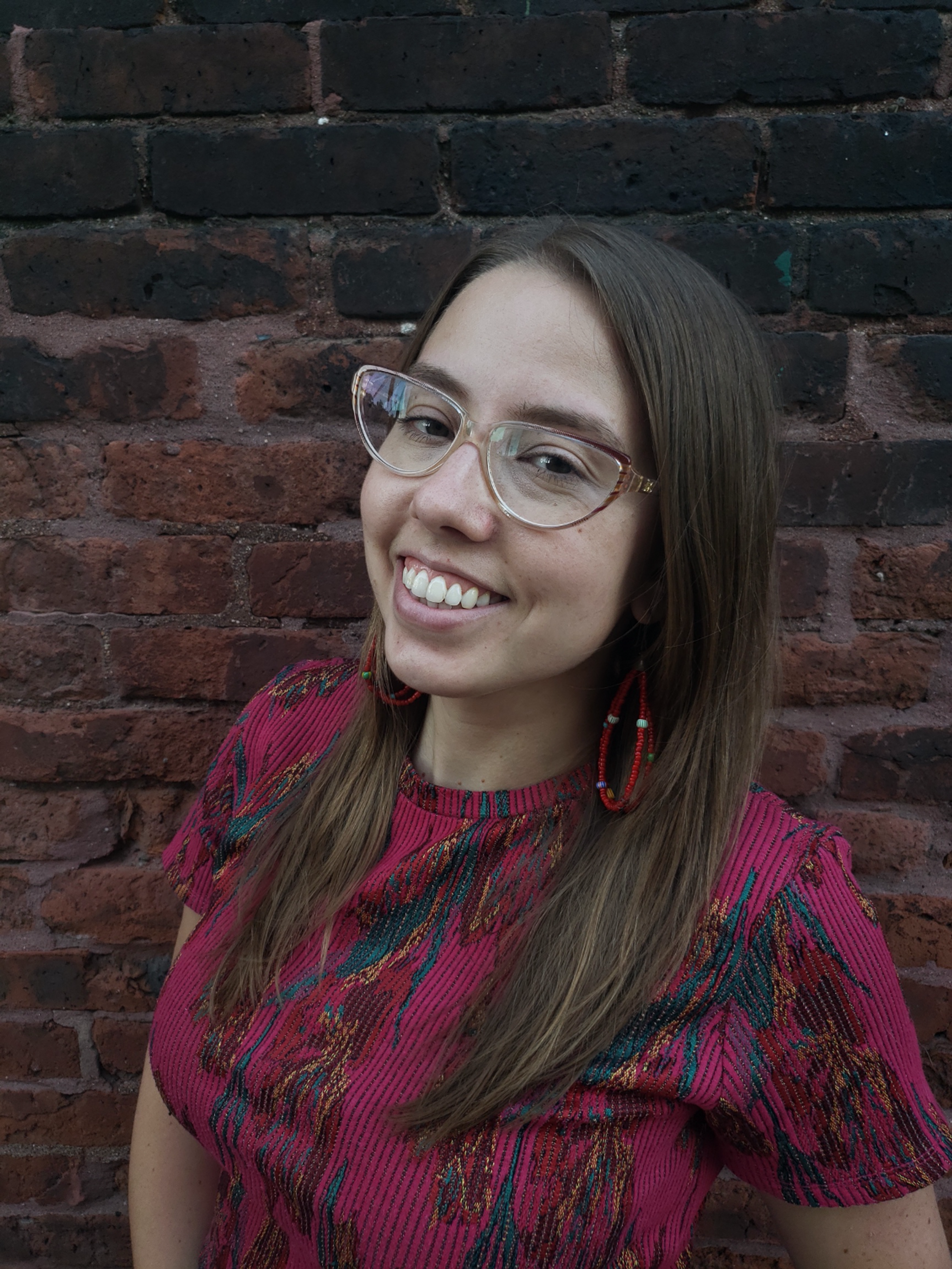 A photo of Carmen Hermo standing against a dark-orange brick wall. Hermo wears glasses and is smiling at the camera.  