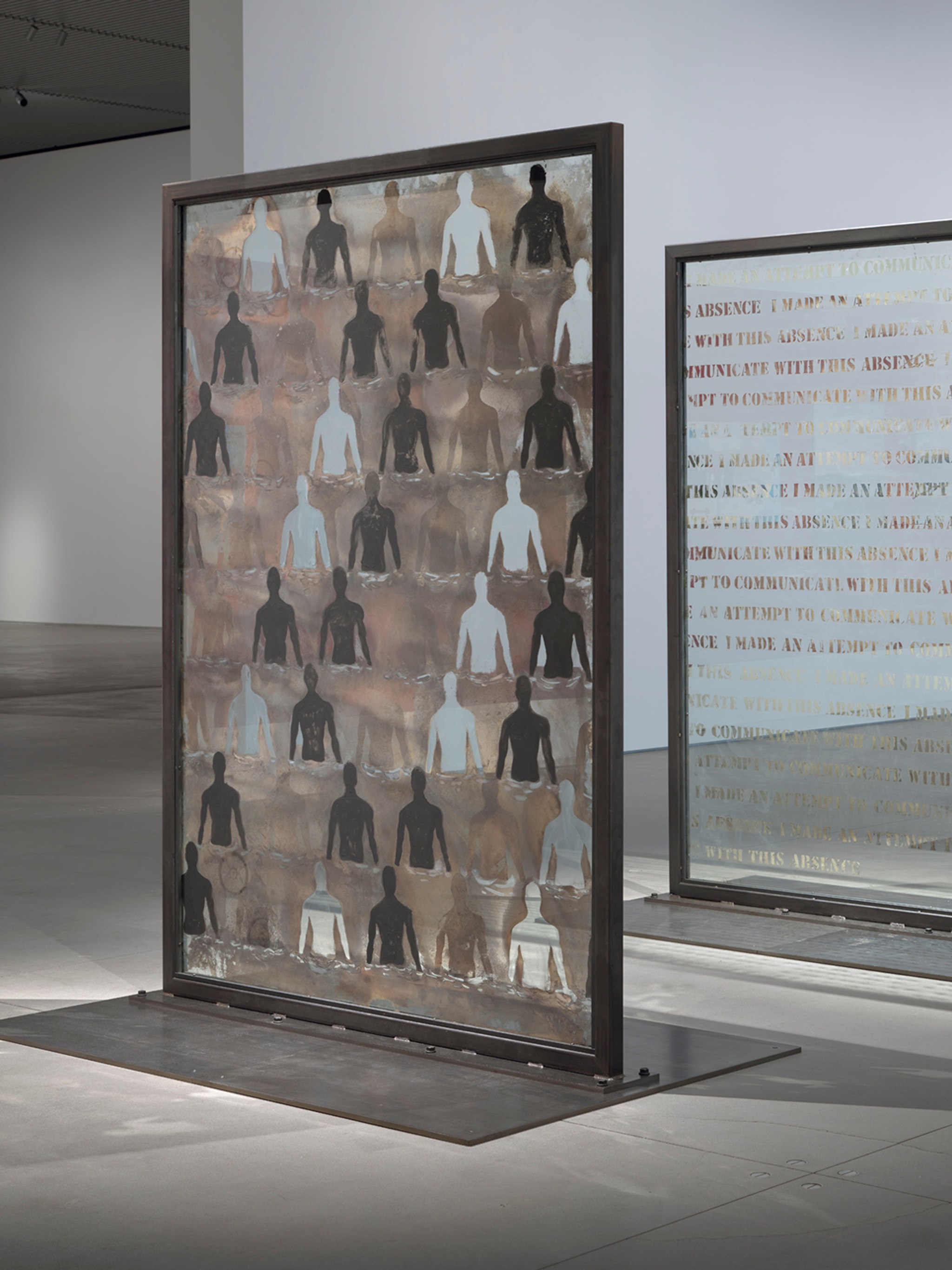 Two glass panels standing upright in a gallery. One has the form of a person from the waist up stenciled in a repeating pattern of mirrored, painted, and transparent glass. The other repeats the sentence "I made an attempt to communicate with this absence.”