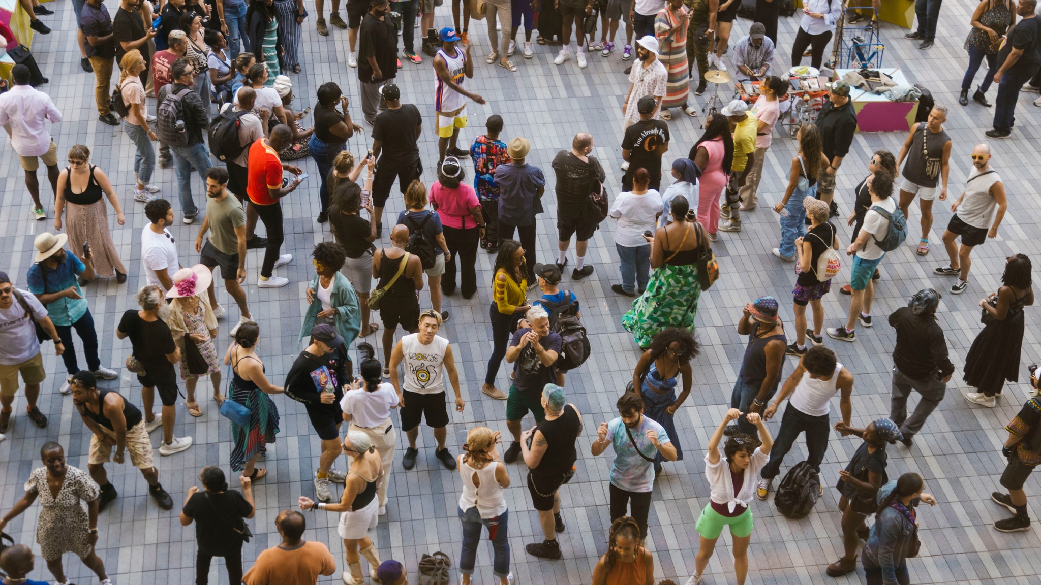 A crowd is seen from above on an outdoor patio with gray paving stones. People are dressed in colorful tshirts and are dancing. One audience member looks up at us. 