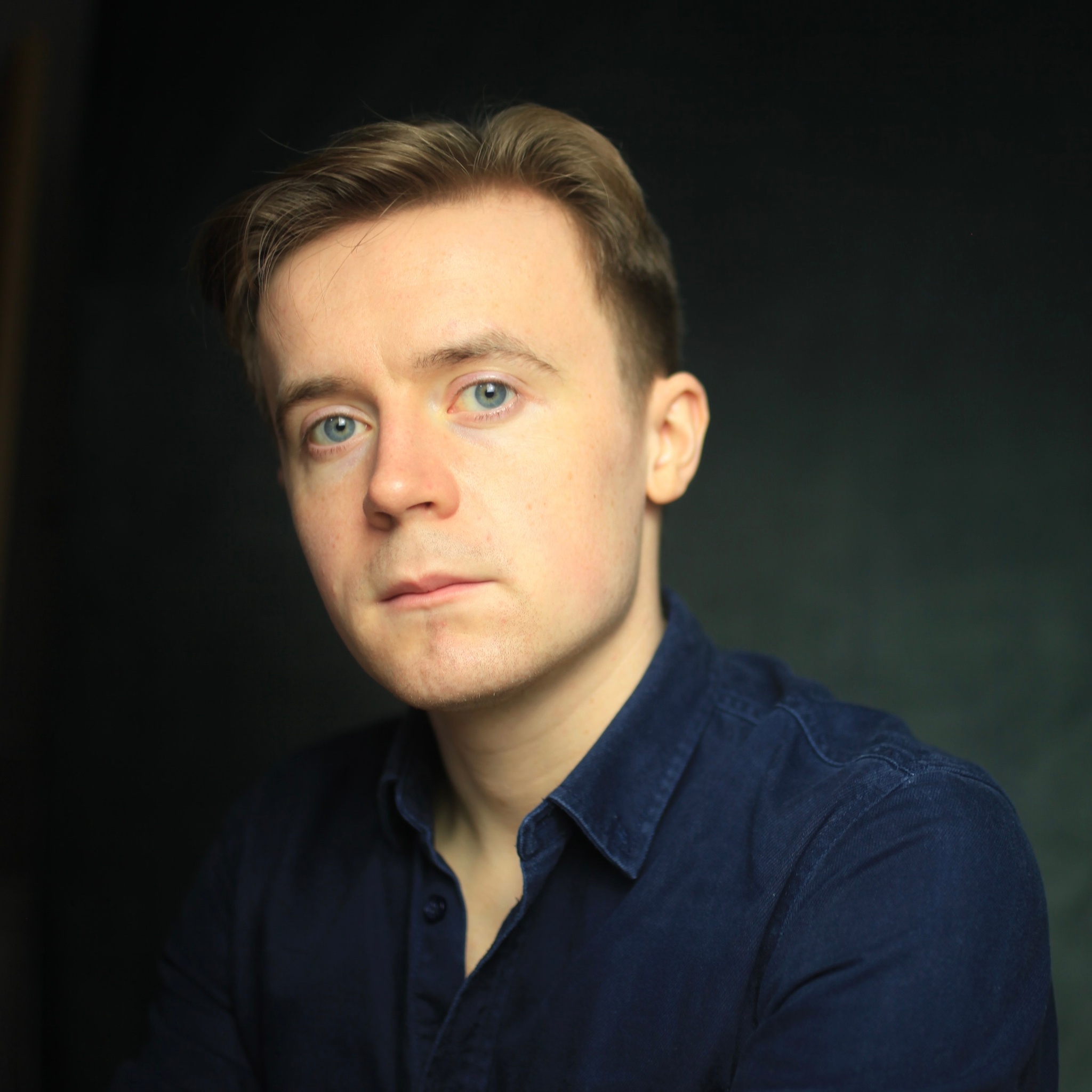 A portrait of actor Stefan Brennan-Healy, a white man with short light brown hair who looks directly at us while cocking his head to the side slightly. He wears a dark blue button up shirt. 