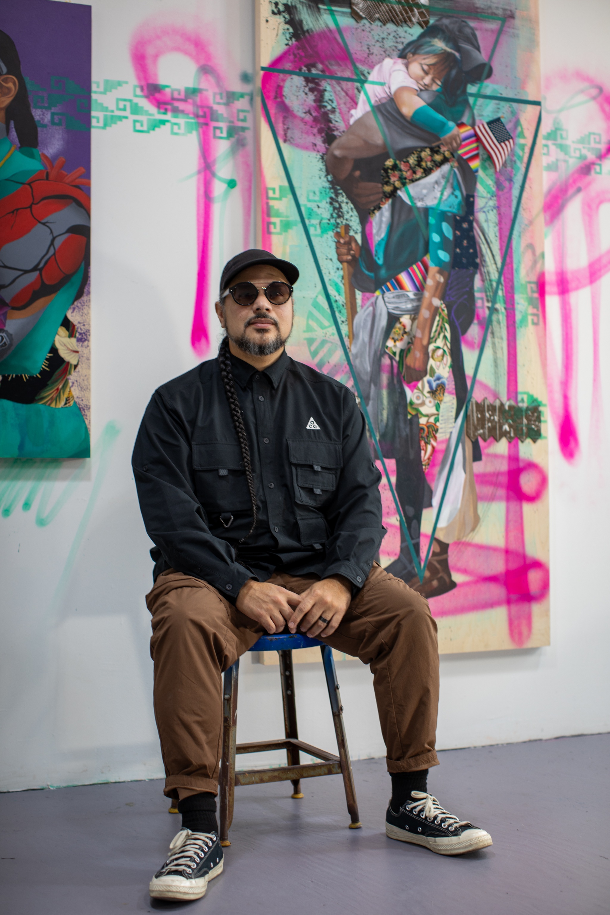 A portrait of artist Victor "MARKA27" Quiñonez. Victor sits in a metal chair in a white studio space. He poses with hands in lap, wearing a cap and dark sunglasses, facing us directly. Behind, colorful graffiti-style paintings hang on the wall. 