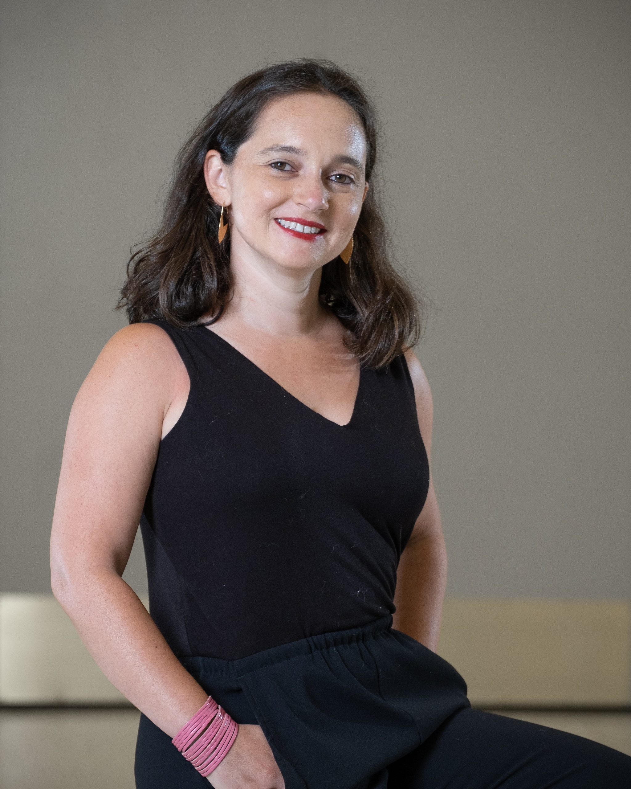 A portrait of Susanna V Temkin, a white woman who poses against a gray backdrop with her hands in the pockets of a black jumpsuit she's wearing. Her brown hair falls to her shoulders. She smiles at us. Photo by Michael Palma Mir