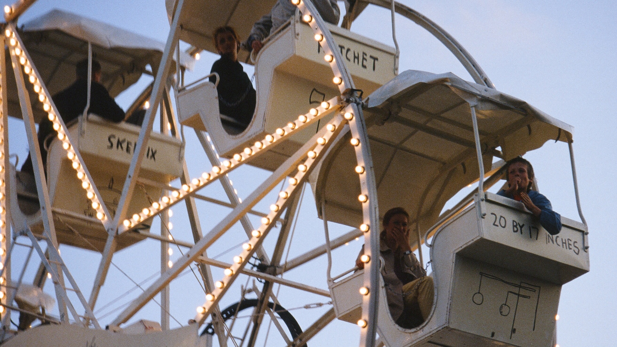 A Ferris wheel gondola with two people inside, seen from below. The Ferris wheel, designed by artist Jean-Michel Basquiat, is white with its scaffolding lined in bright white light bulbs. 