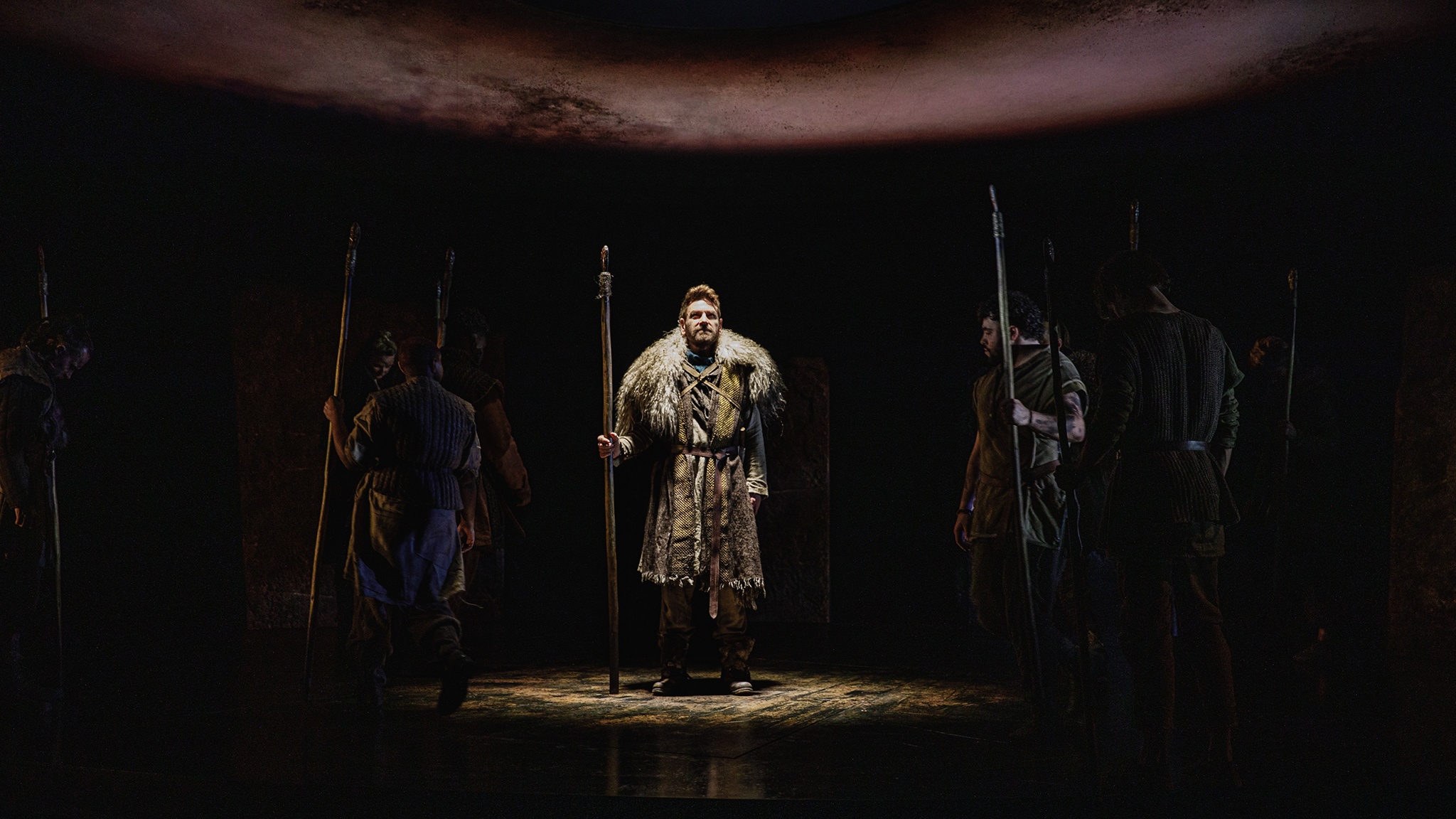 A production photo of Kenneth Branagh on stage as King Lear. He stands at the center of the circular stage in a costume including a fur pelt draped over his shoulders. His is lit in a focused light while around him are other members in the cast in shadow. 