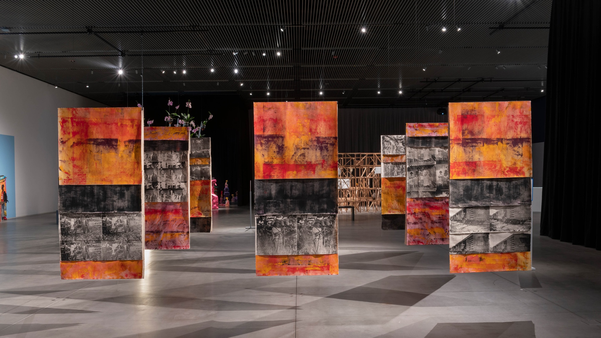 Nine painted fabric panels hang from a gallery ceiling in a three by three grid. The panels are painted in orange and black bands, with screen printed sections with black and white images of banana plantation workers in the 1920s. 