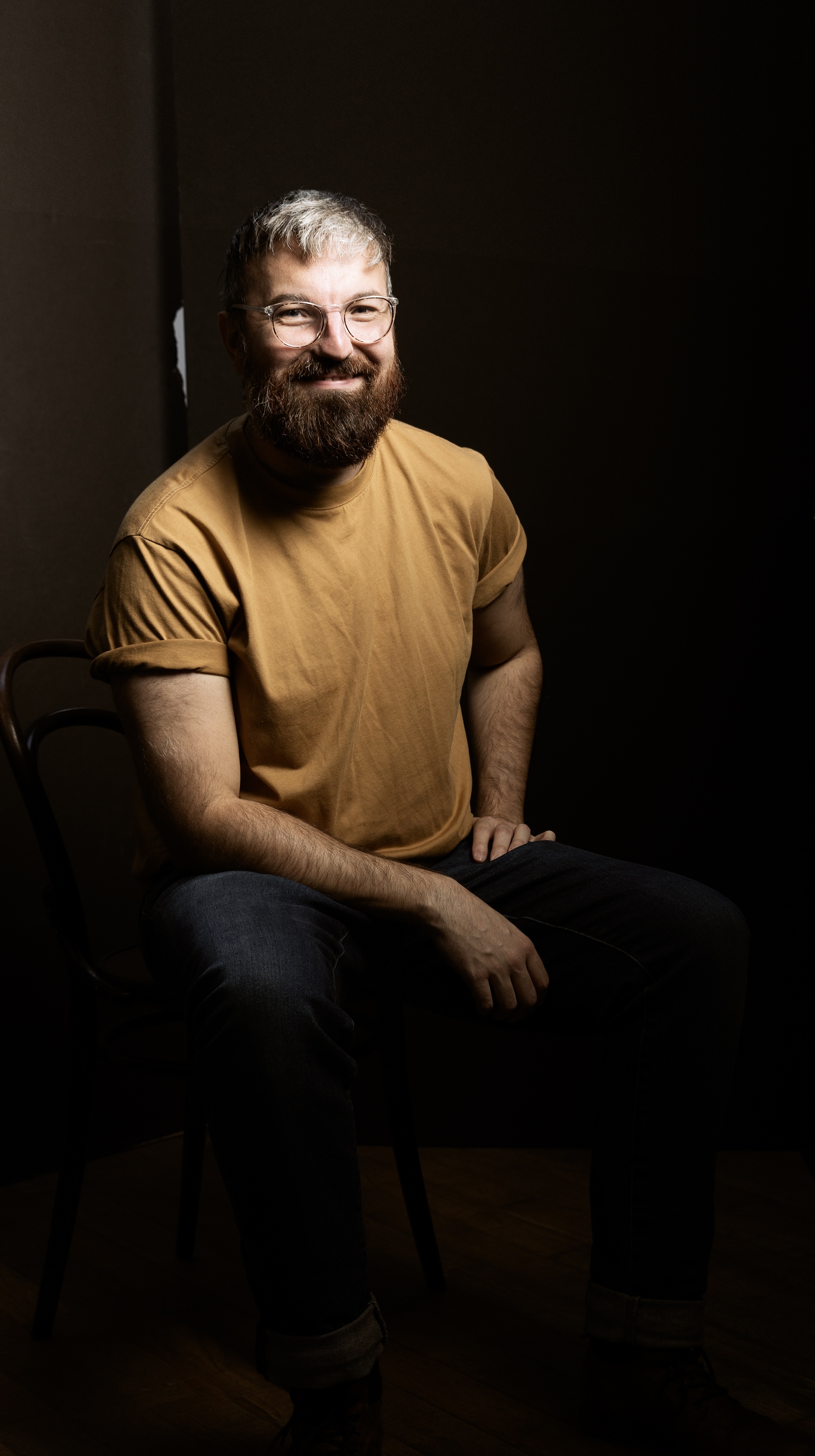 A portrait of artist Andrew Morrill. He is a white man who is pictured in dramatic lighting with the background in shadow. He leans slightly forward on one arm propped on his leg. He had a beard, wears glasses and a golden brown t-shirt, and smiles at us. 