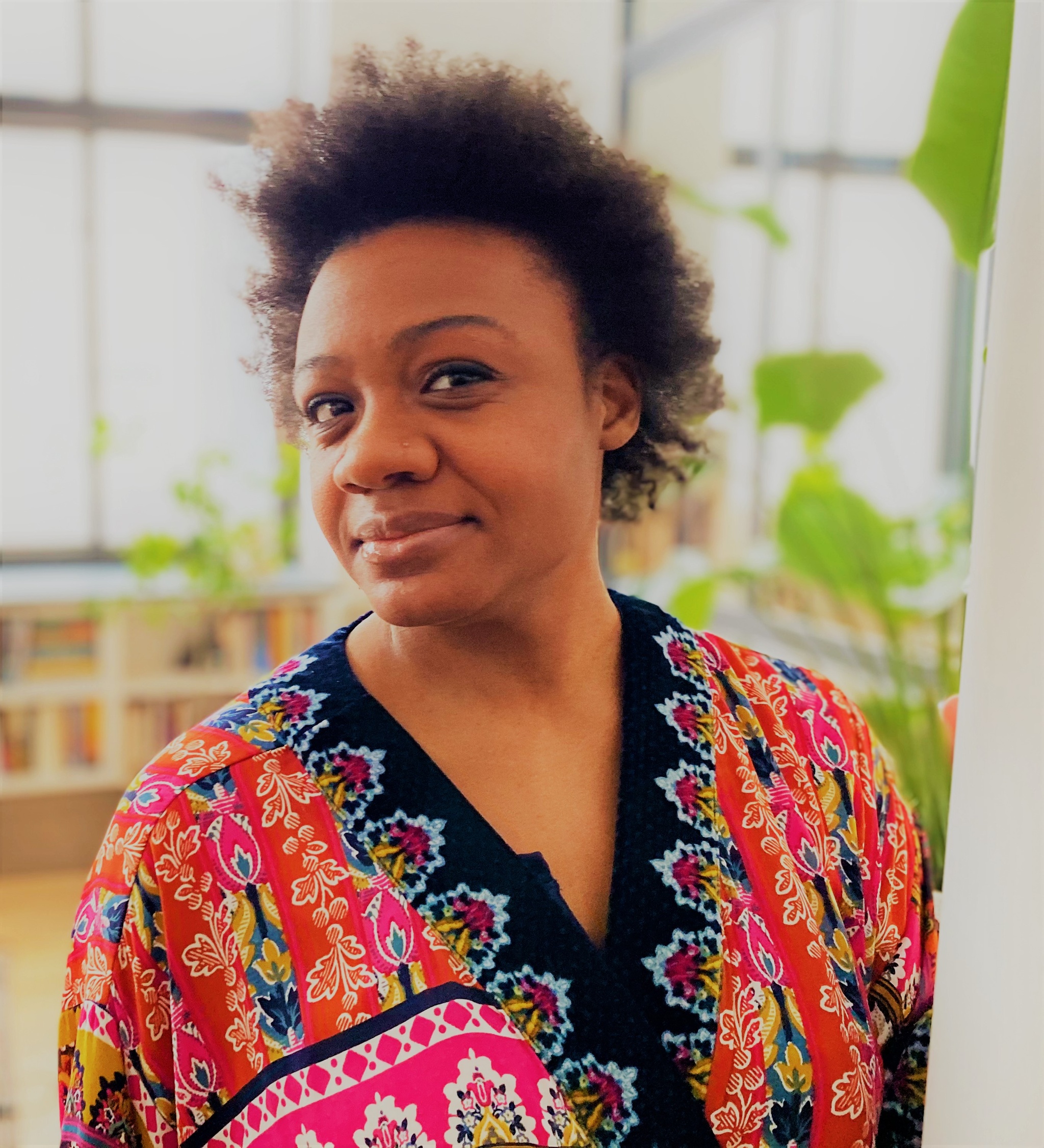 A portrait of Rakia Seaborn, a Black woman who wears a colorful printed v-neck blouse and raises one eyebrow at us playfully.