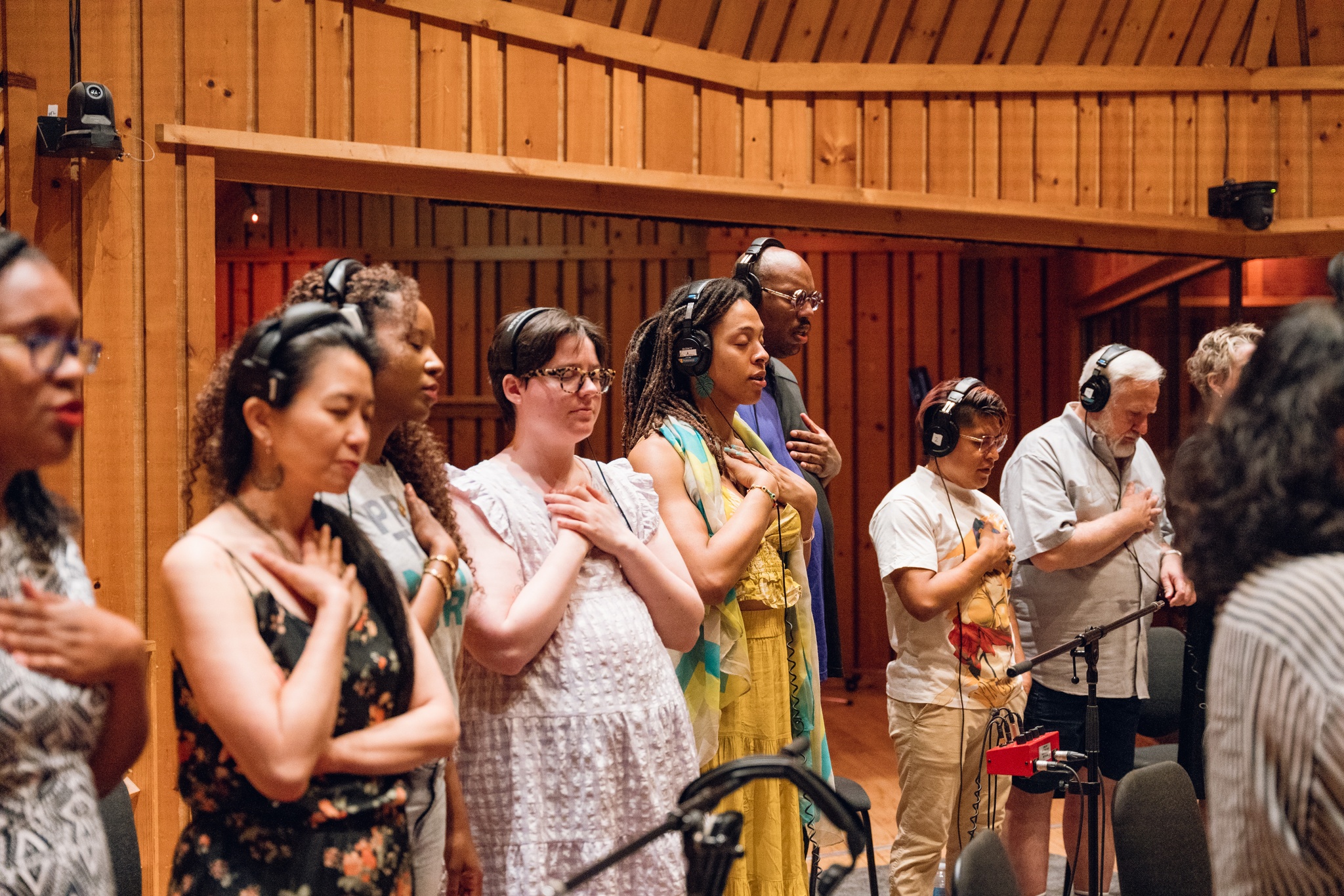 Choir members gathered in the recording studio stand in a row with eyes closed, heads slightly bowed, and hands crossed over their hearts. 