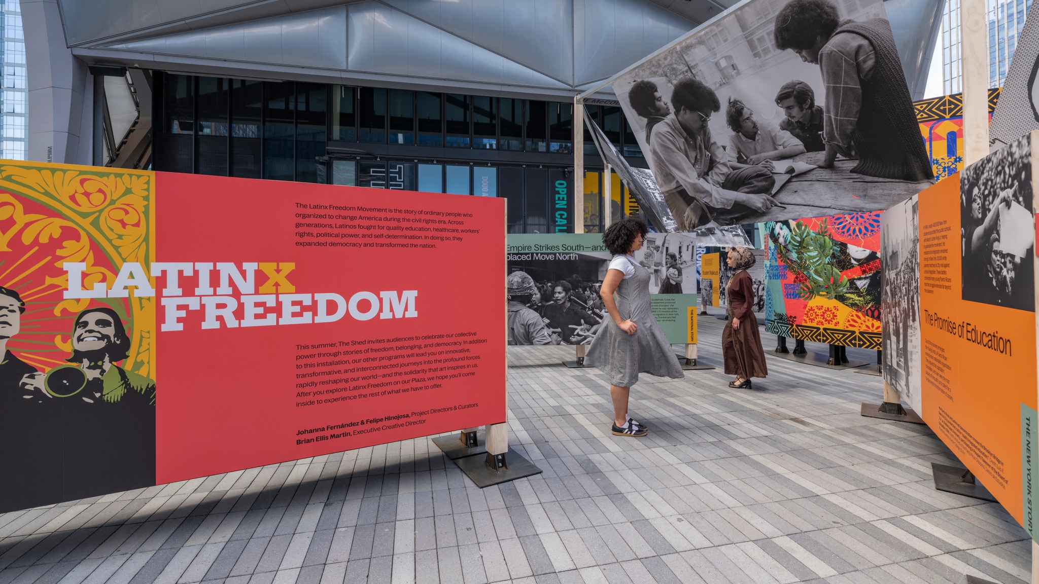 An outdoor art installation displays historical photos from the Latinx civil rights movement. Two exhibition-goers stand among large panels displaying the photos. Overhead a banner with a black and white photo of activists billows in a breeze. 