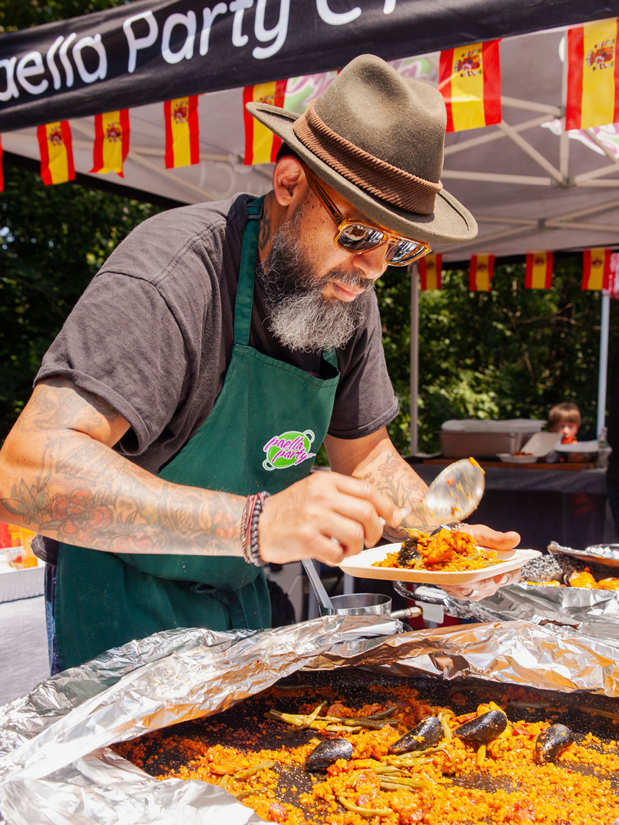 A man in a fedora hunches over a large pan of paella in the sun. He scoops some of the rice onto a plate. 